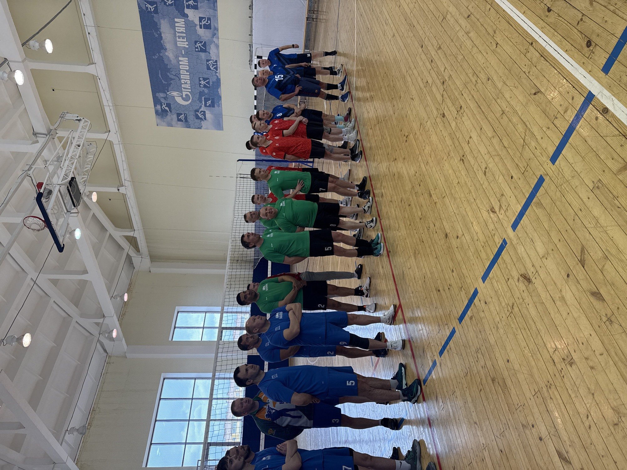 The image depicts an indoor sports hall in Noviy, Russia, during what appears to be daytime. Approximately 20 to 25 adult males are lined up on a wooden sports court, arranged in several rows. Most are wearing athletic attire, including shorts and t-shirts or jerseys in blue, green, and red colors, suggesting different teams. Numbers are visible on some jerseys, such as 15, 8, 7, 5, 2, 3, 4, and 13.
The men are standing upright, many with arms crossed, in a formal lineup, possibly before or after a volleyball match or training session, given the presence of a volleyball net stretched across the court. The floor is made of light-colored wooden planks, marked with blue and white lines typical of a sports facility.
The hall is illuminated by bright overhead lights, and large windows are visible high on the walls, allowing natural light to enter. A prominent blue banner hangs on one wall, displaying the Cyrillic text "ГАЗПРОМ - ДЕТЯМ," which translates to "GAZPROM - FOR CHILDREN," alongside various pictograms depicting sports activities. A portion of a basketball hoop structure is also visible on the left side of the frame.