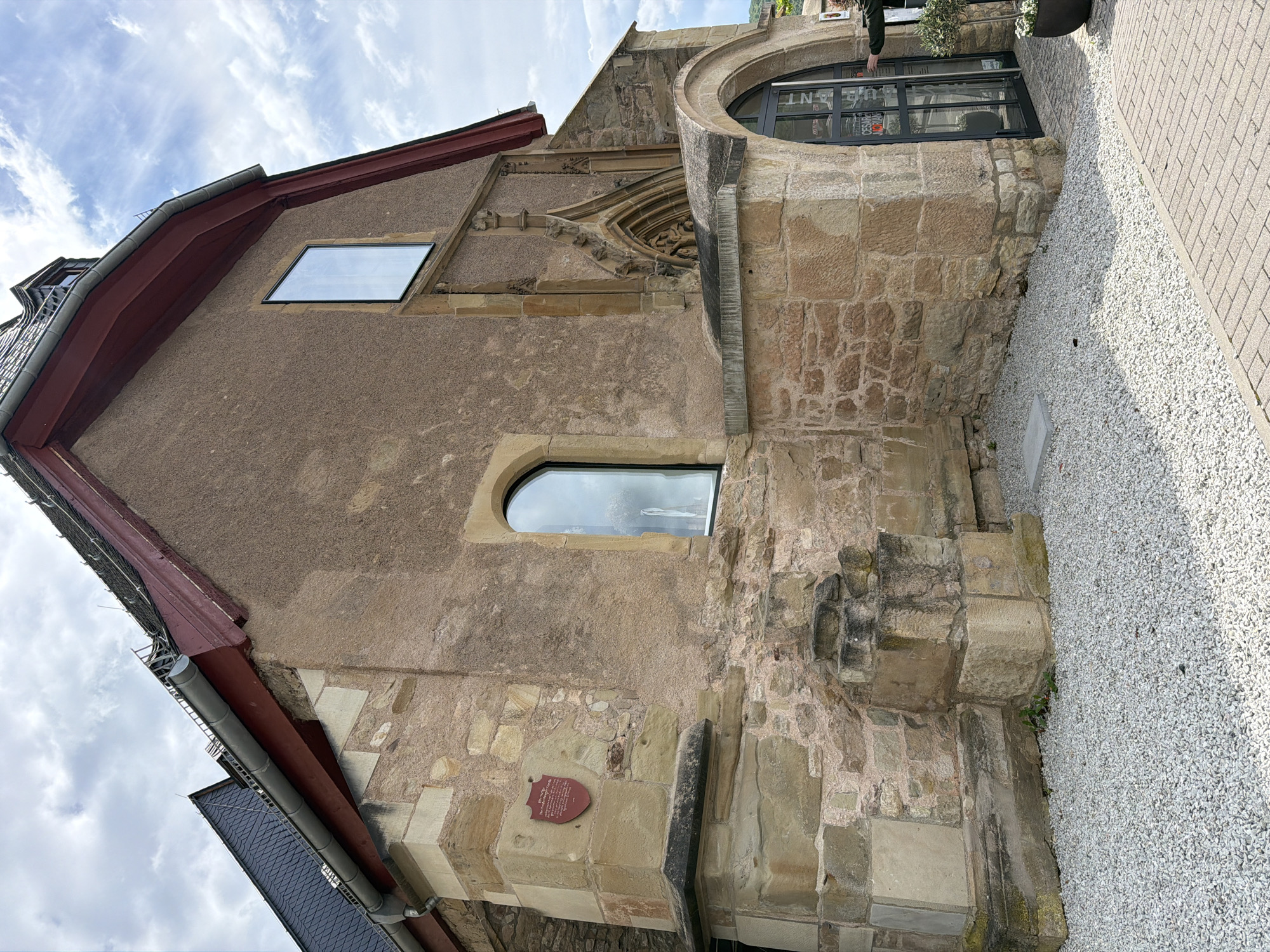 The photograph depicts the exterior of the Synagogue Bad Sobernheim, located in Bad Sobernheim, Germany. The structure, viewed from a low angle, features a blend of architectural styles, with a mix of stone and stucco facades. It incorporates both historical and modern elements. A large, rounded window is integrated into a stone section of the building. Two additional windows, one rectangular and the other arched, are visible on the building's side. A plaque is mounted on the building's facade. The surrounding ground is covered in gravel, and a paved sidewalk borders the building. The sky above is partly cloudy.