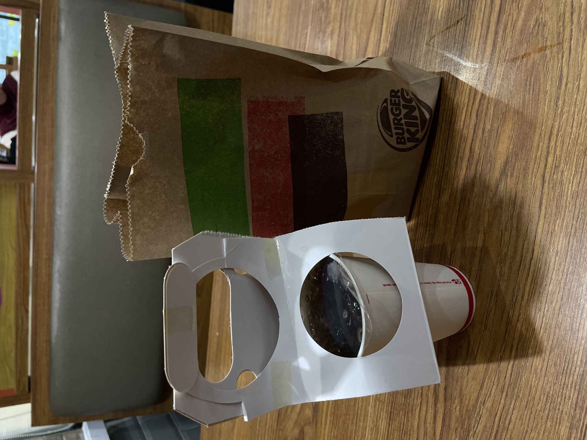 The image shows a Burger King paper bag and a drink carrier with a cup on a wooden table. The Burger King bag is brown and has the company's logo printed on it in white. It also features three colored stripes: green, red, and brown. The drink carrier is white and has two circular cutouts, one of which holds a white cup with red accents and black liquid inside. The cup has some red text on it, but it is difficult to read.
The scene appears to be an indoor setting, possibly a fast-food restaurant or a home, given the wooden table surface. There are no people prominently featured in the image, although a blurred figure can be seen in the reflection of a framed object in the background. The lighting suggests it is daytime. The overall impression is a casual moment, perhaps after a meal or a takeaway purchase. The location context of Davao City, Philippines, is provided, but there are no specific visual cues in the image that directly confirm this location.