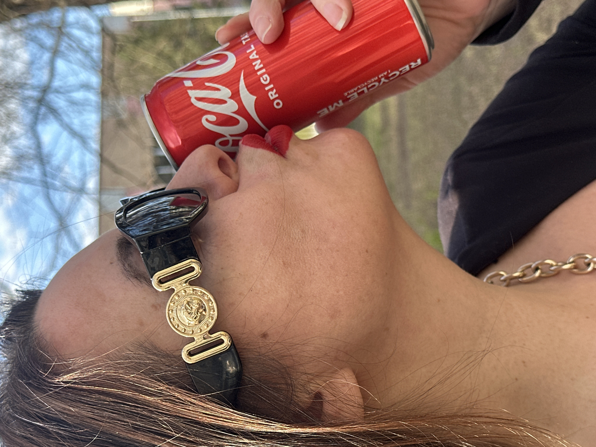 A woman is drinking from a red Coca-Cola can. The can is tilted at an angle towards her red lips. She is wearing a black and gold sunglasses on her forehead and a gold chain around her neck. The background appears to be outdoors, possibly a park with trees, but the location is not possible to be identified as Riga, Latvia from the visual data provided.