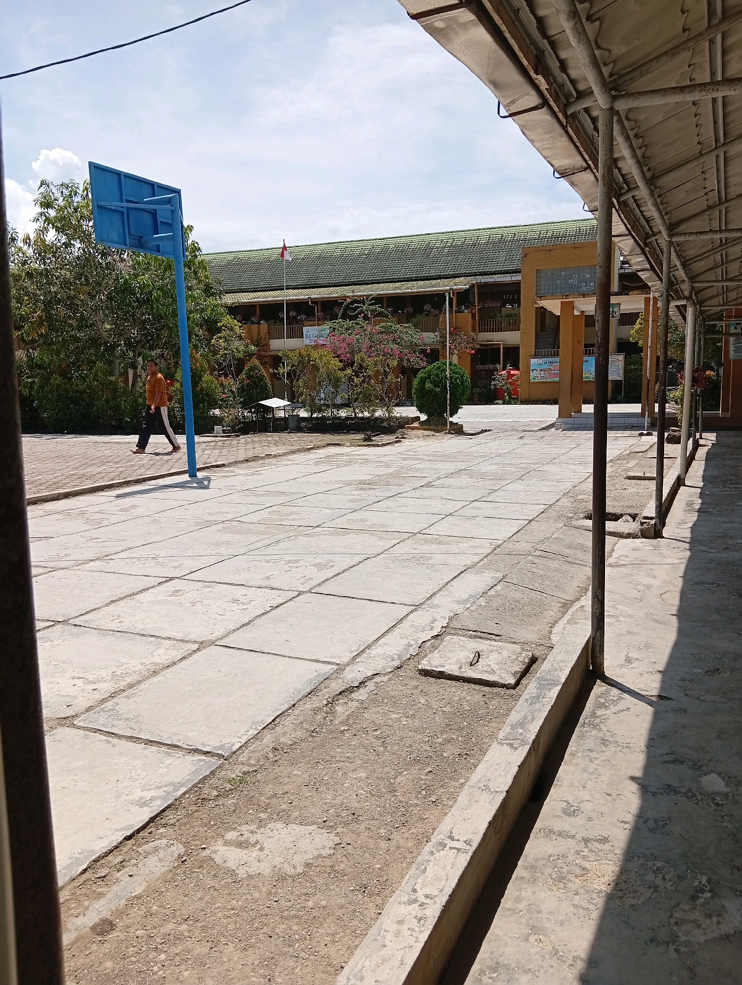 The image shows an outdoor scene in the City of Gunungsitoli, Indonesia, likely a schoolyard. In the foreground, a paved area with square concrete tiles is visible, bordered by a concrete drainage channel and a covered walkway with metal support poles. A person wearing an orange shirt and dark pants walks across the tiled area towards the left. A blue basketball hoop stands on a pole to the left of the frame. In the background, a building with a green tiled roof and multiple stories is visible, featuring balconies and various decorations. A red and white flag is planted on a pole in front of the building. Trees and flowering bushes are scattered throughout the scene, adding greenery and color. The sky is clear with some scattered white clouds. The lighting suggests it is daytime, possibly mid-morning or afternoon, given the distinct shadows cast by the walkway's structure.