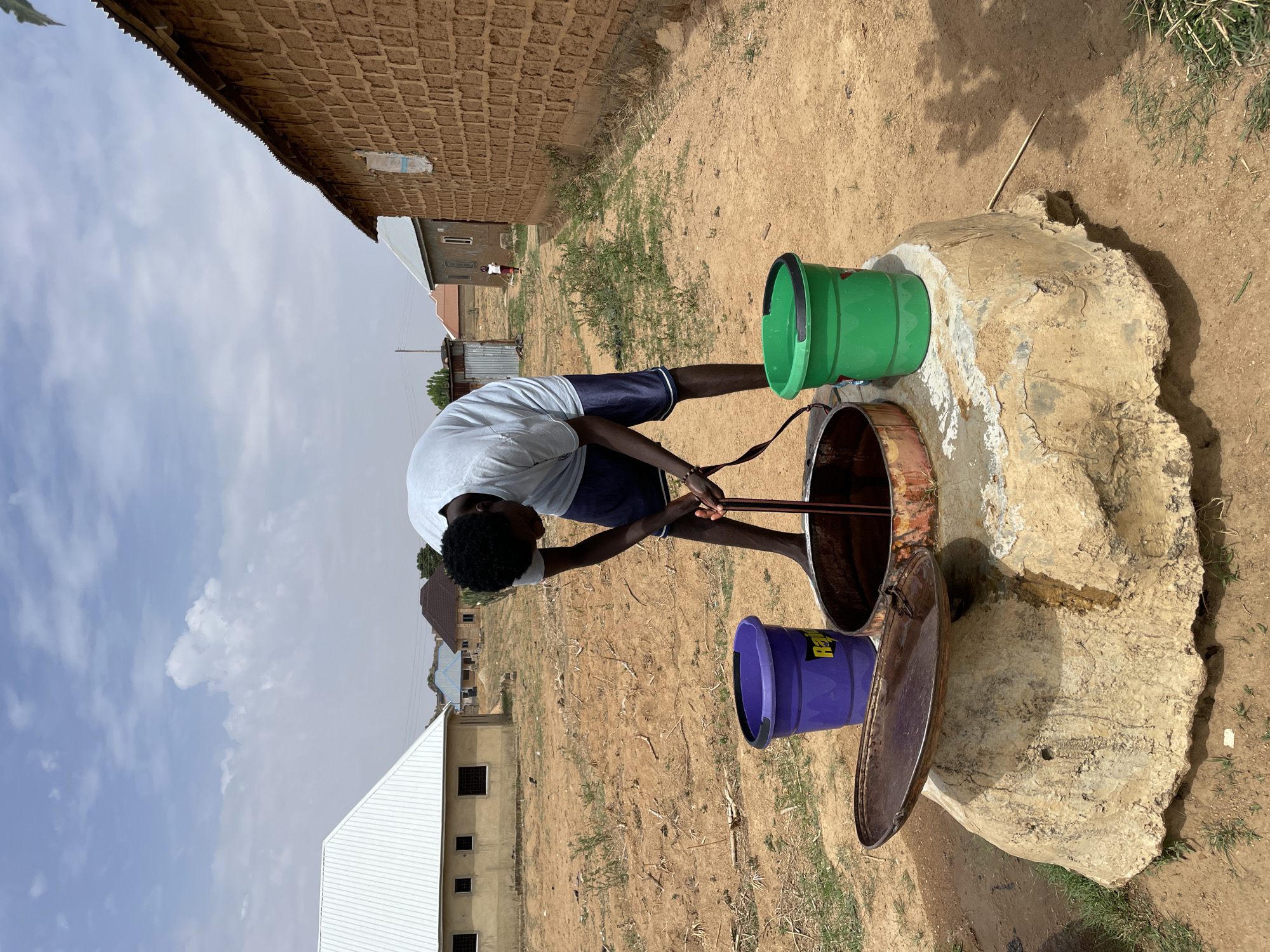 In a daytime, outdoor scene in Jos, Nigeria, a person is drawing water from a well. The person is of African descent, wearing a light-colored t-shirt and dark shorts. They are bending over the well, using what appears to be a rope or chain to lower a bucket into it. Two buckets, one green and one purple, are positioned on the well's rim. The well is circular with a rusted metal lining and is set in a large, roughly hewn stone or concrete base. The surrounding environment is dry and dusty, with sparse vegetation. Several buildings are visible in the background, including some with corrugated metal roofs and brick structures. The sky is partly cloudy.