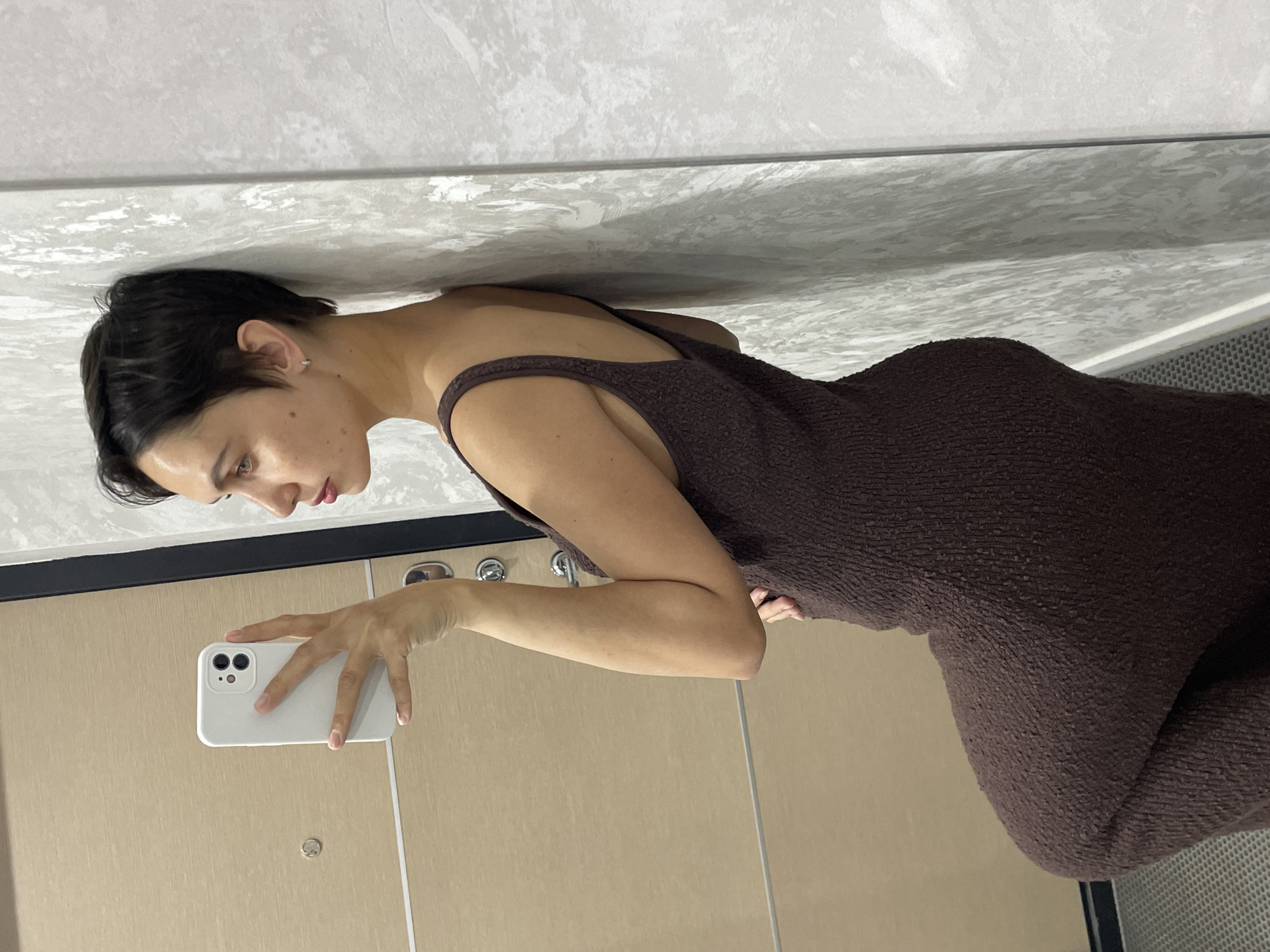 A woman is in a bathroom, facing a mirror and holding a white cell phone in her right hand, presumably taking a selfie. The bathroom has light-colored, smooth walls and a light-colored textured ceiling. The woman is wearing a brown, possibly textured, sleeveless dress. Her head is tilted downwards as she looks at the phone screen. Her dark hair is short. The location in Izhevsk, Russia cannot be determined.