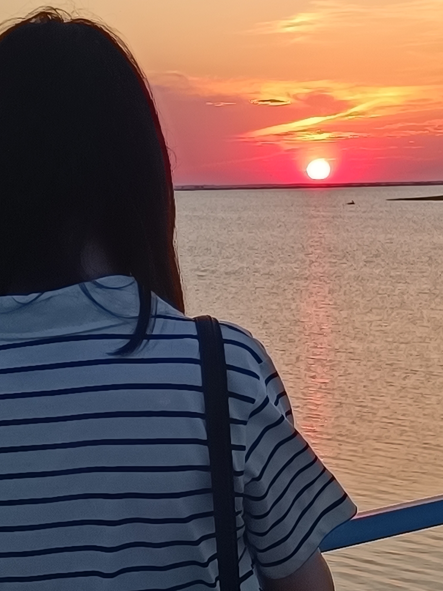 A person stands facing a body of water, their back to the viewer. The person has dark hair and wears a striped, collared shirt with a dark strap over their shoulder. The setting appears to be at sunset, with a red sun low on the horizon, reflected in the water. Based on the presence of the lake, this image was likely taken at Lake Kopa in Kokshetau, Kazakhstan.