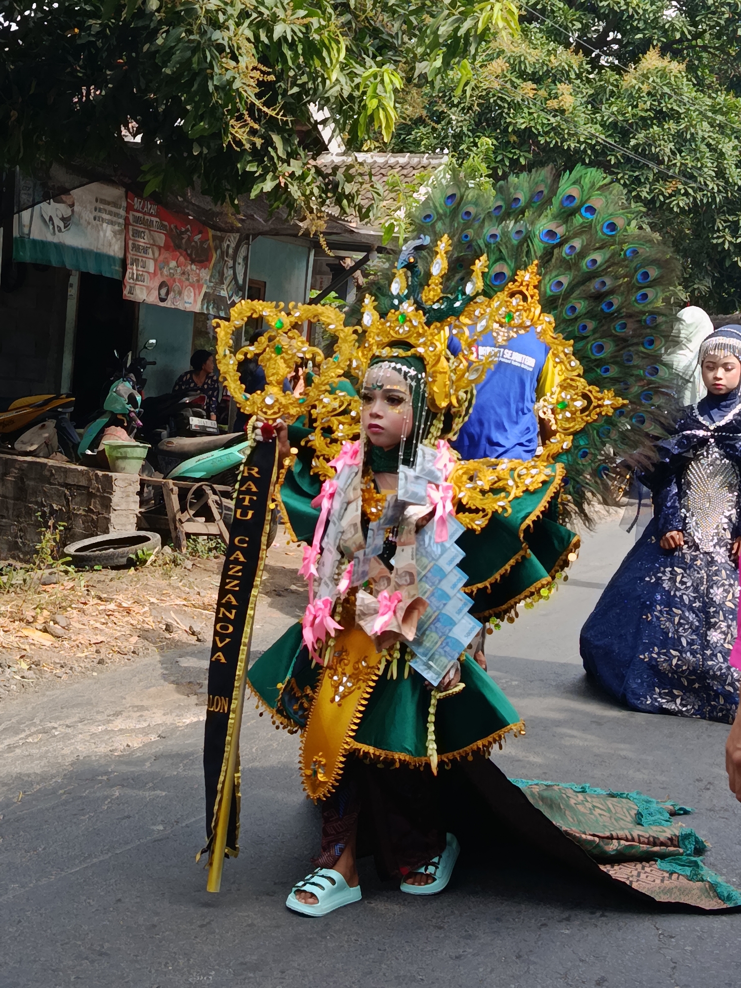 In Ketowan, Indonesia, on a sunlit day, a young person stands center-frame, adorned in an elaborate peacock-themed costume. The costume features a large, fanned tail of iridescent peacock feathers, a golden, ornate headpiece, and a green, layered garment. Strands of Indonesian Rupiah banknotes are draped around the neck, intertwined with pink ribbons and yellow embellishments. A black banner with gold lettering, reading "RATU CANZANOVA," hangs from the costume. The individual wears light blue sandals.
To the right of the main subject, another young person, dressed in a dark blue, sequined hijab and dress, observes the scene. Behind them, a building with a red and white banner, possibly advertising a business with images of motorcycles and a car, is visible. Several motorcycles are parked near the building. The ground is paved, with dry leaves and debris scattered around. Trees with lush green foliage provide shade in the background. The overall atmosphere suggests a local festival or parade.