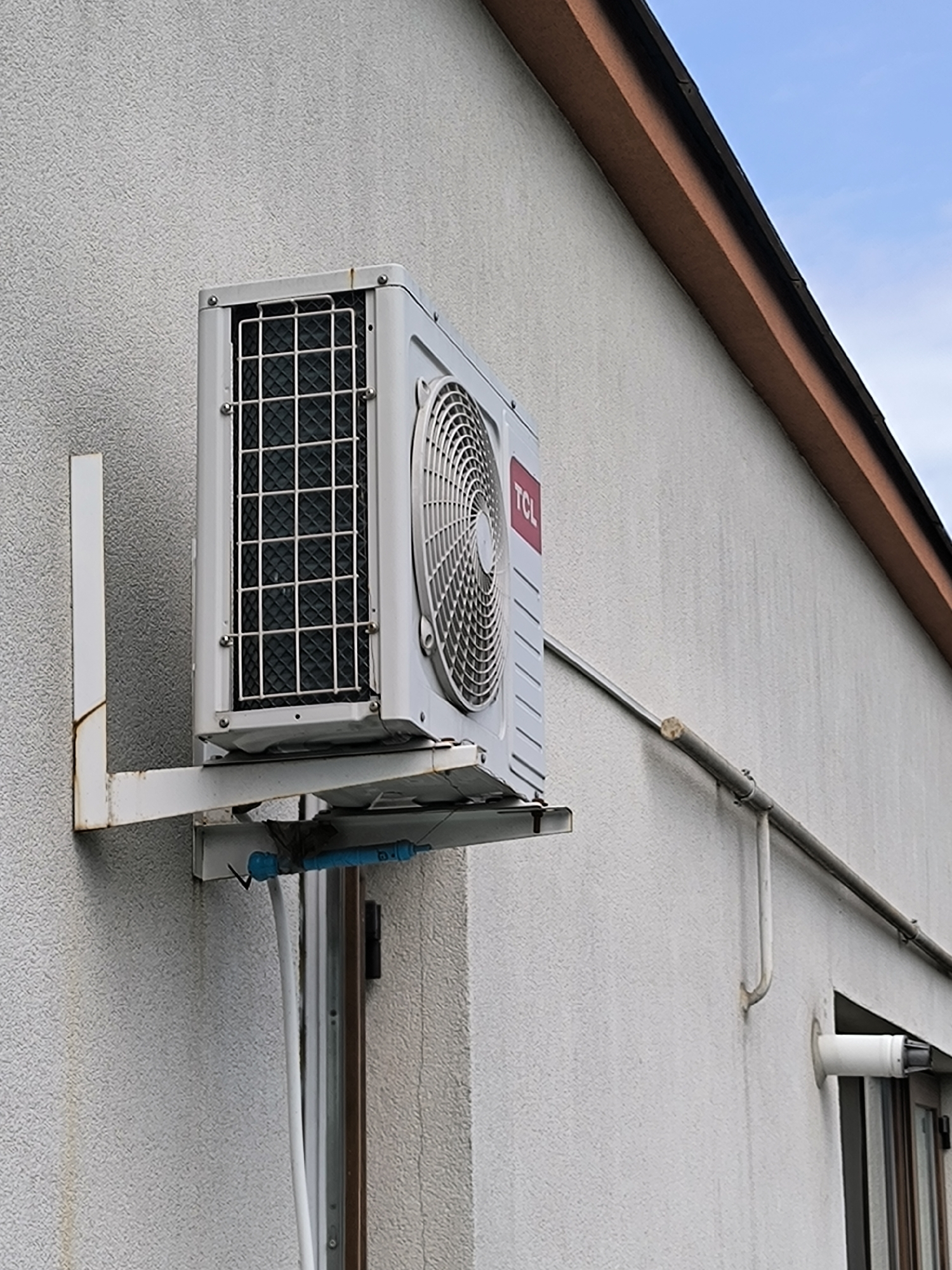 An outdoor air conditioning unit, branded TCL, is mounted on a textured white exterior wall of a building. The unit is white and has a visible fan grille and an inlet grille covered with a wire mesh. It is supported by a white metal bracket attached to the wall. Below the unit, blue pipes and a white conduit are visible. A metal pipe runs horizontally along the wall to the right of the unit. In the upper right portion of the image, a segment of a brown soffit is visible against a pale blue sky with some clouds. The scene appears to be an outdoor residential or commercial setting in Chișinău, Moldova, as indicated by the architectural style and typical placement of such units. There are no people or animals present in the image.