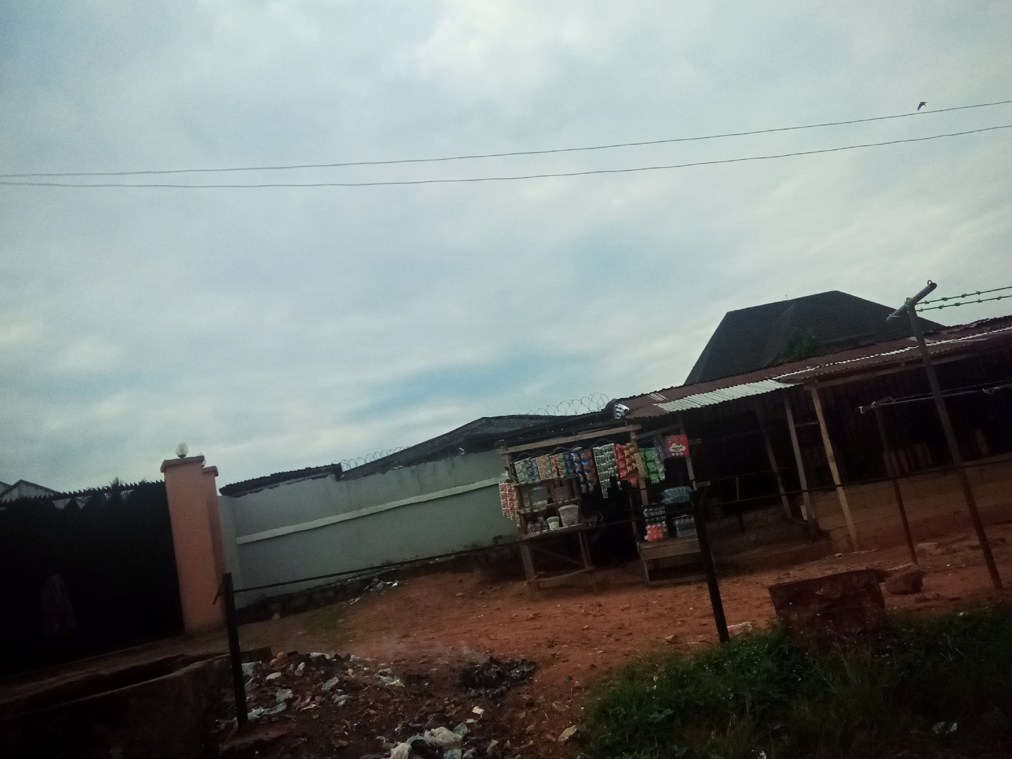 The image depicts a scene in Oko Town, Nigeria, on an overcast day. In the foreground, a dirt path is visible, littered with refuse and debris, including plastic bottles and what appears to be burnt material. To the left, a segment of a tall, peach-colored pillar with a decorative top is seen, adjacent to a dark, wrought-iron gate. Beyond the gate, a structure with a dark, pointed roof is partially visible.
Dominating the midground is a roadside stall, constructed with wooden planks and a corrugated metal roof. The stall displays various packaged goods, including drinks and snacks, arranged on shelves. Barbed wire is strung along the top of a low, grey concrete wall that runs behind the stall, extending to the left. The wall appears to be a boundary.
In the background, the sky is cloudy and muted in color, with two parallel power lines stretching across it. A small bird is captured in flight high in the sky. The overall impression is one of a utilitarian, outdoor commercial space within a residential or semi-urban setting.