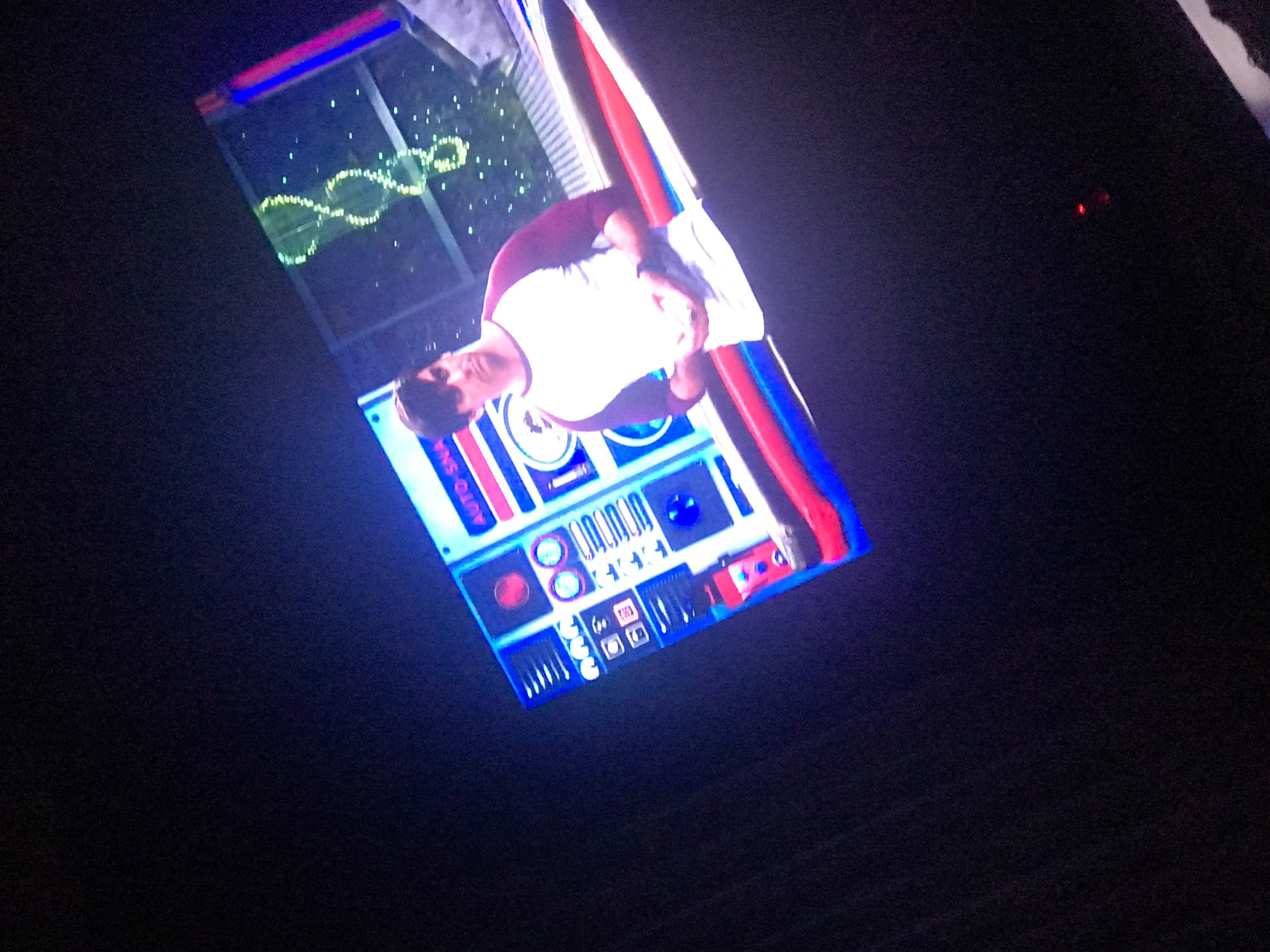 The image shows a man standing in front of an interactive display, presumably a museum exhibit or art installation. The display has various buttons, lights, and symbols, along with the words "AUTO SNAX". Above the man and the display is a window or screen with a lit-up DNA-like structure on it. The overall color scheme is a mix of blues, reds, and whites, creating a neon-lit effect. Based on the provided information, this is located in the city of Yerevan, Armenia. The man is wearing a white shirt with maroon trim.
