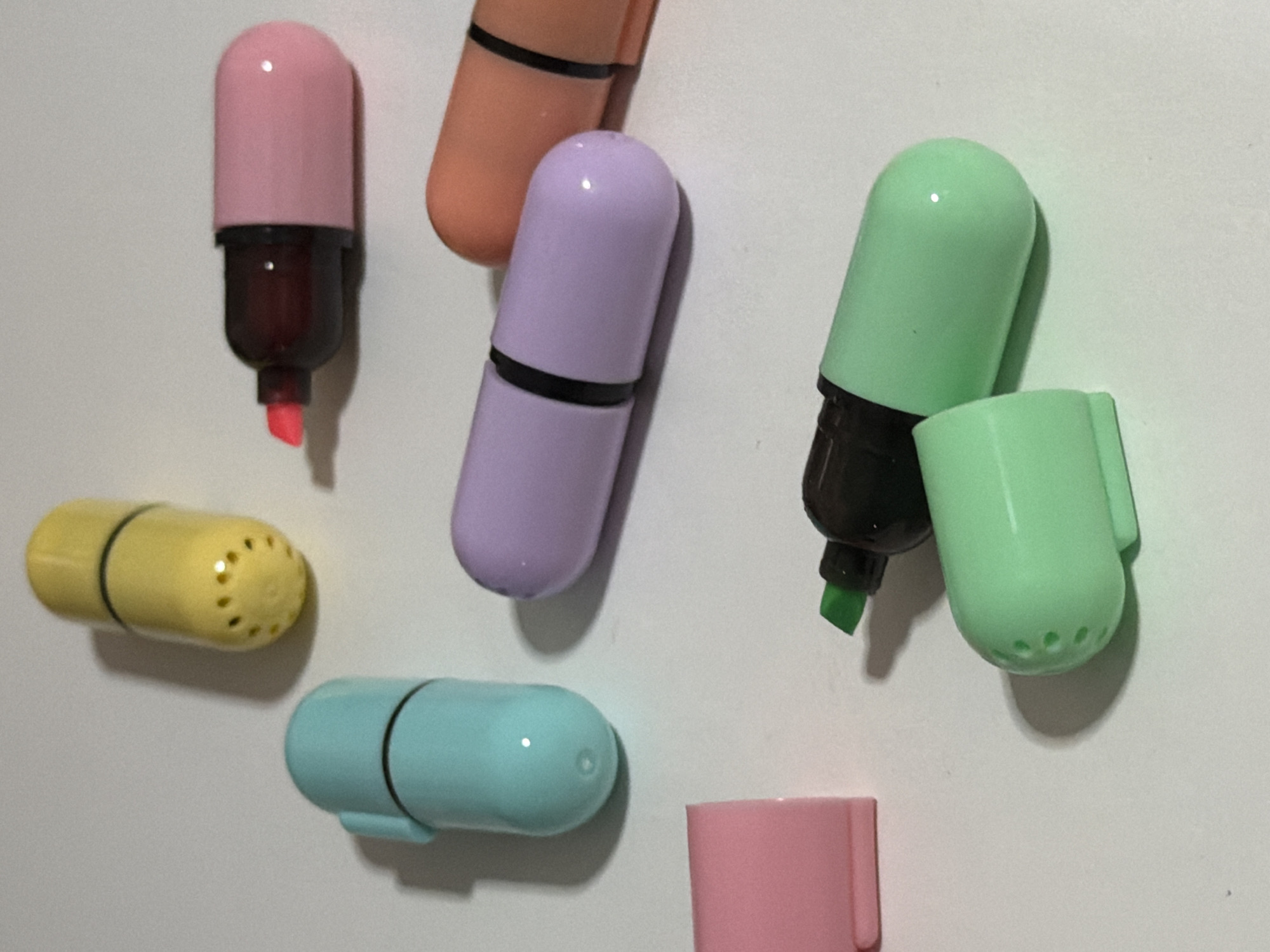 A top-down shot shows a collection of pastel-colored capsule-shaped highlighters on a plain white surface. There are seven highlighters in total, some with their caps on and some with their caps off. From left to right, top to bottom, a pink cap is visible. Next to it, a yellow highlighter with its cap on, showing small ventilation holes. To its left, a turquoise highlighter with its cap on. Below that, a lavender highlighter with its cap on. To the right of the lavender highlighter, an orange highlighter with its cap on. In the foreground, a green highlighter with its cap off, revealing a green felt tip. Next to the green highlighter, its cap lies open. The lighting is bright, creating subtle shadows that indicate the objects are placed on a flat surface. There is no indication of location within Moscow, Russia.