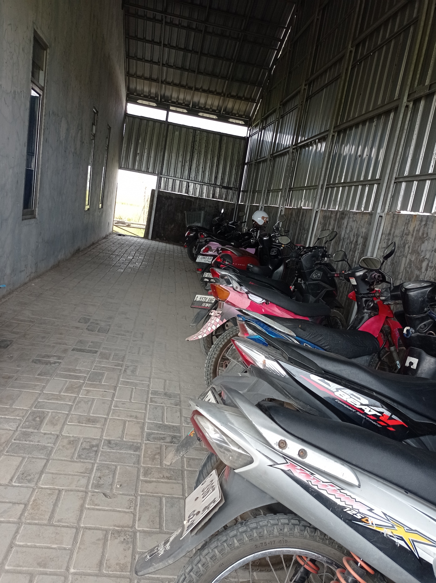 The image depicts an outdoor, covered parking area, likely a carport or a section of a building, filled with a row of parked motorcycles. The floor is paved with interlocking grey concrete bricks. The structure has a metal roof supported by beams, and the walls appear to be constructed from concrete on the left and corrugated metal siding on the right. Natural light enters from an open end at the back of the structure, illuminating the scene. Several motorcycles are visible, primarily in shades of red, black, and silver, with some showing colorful decals. License plates are visible on some of the motorcycles, with one reading "C 2963 AJF" and another "C 5374 BHG". A white helmet is placed on one of the motorcycles. The scene suggests a practical, utilitarian space for motorcycle storage. The location is identified as Maribaya, Indonesia.