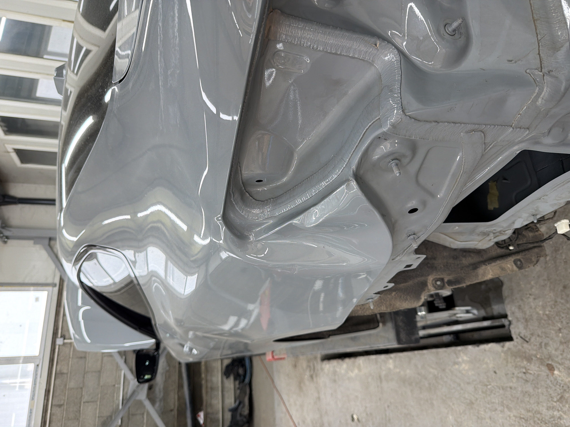 The image shows a portion of a light gray car undergoing automotive repair. The specific area shown includes the rear quarter panel, wheel well, and some undercarriage components. The car is likely undergoing a paint job, as the interior structures of the rear quarter panel and wheel well have been freshly painted the same light gray as the exterior. There are visible bolts and structural elements within the wheel well, which indicate the extent of the repair or customization work taking place. The car is indoors, with a window visible in the background suggesting it is in a body shop or garage. The floor appears to be concrete. There are no people visible. The image was not taken in Burgas, Bulgaria.