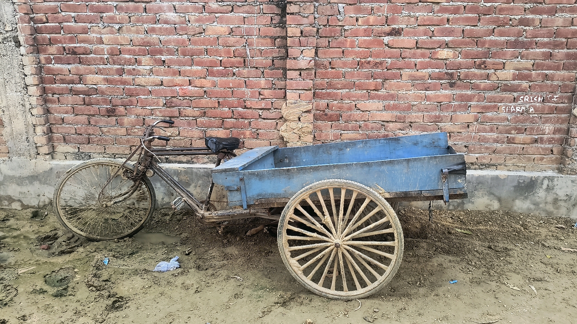A three-wheeled cart, attached to a bicycle, is parked on a muddy ground in front of a red brick wall. The cart is painted blue and has a wooden box on top. The bicycle has a black seat and a chain. The ground is wet and muddy, with some debris scattered around. The brick wall is old and weathered, with some graffiti written on it. The scene appears to be in Loni, India.