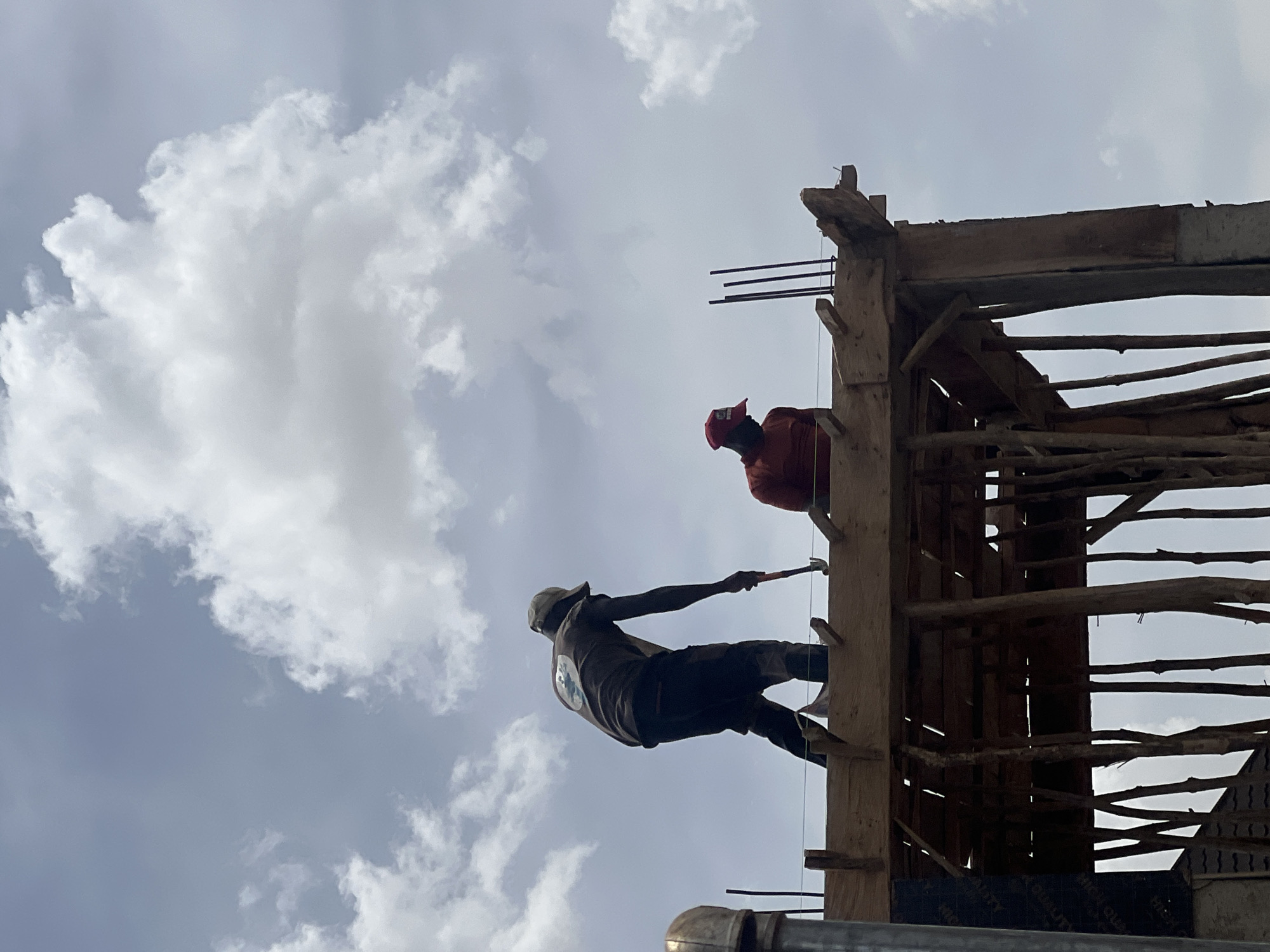 Two men are on a construction site in Jos, Nigeria. One man, wearing a red cap and orange shirt, is standing inside a structure made of wood. The other man, wearing a grey cap and dark shirt, is balanced on a piece of wood while hammering. The sky is cloudy. Steel rebar protrudes from the top of the wooden structure.