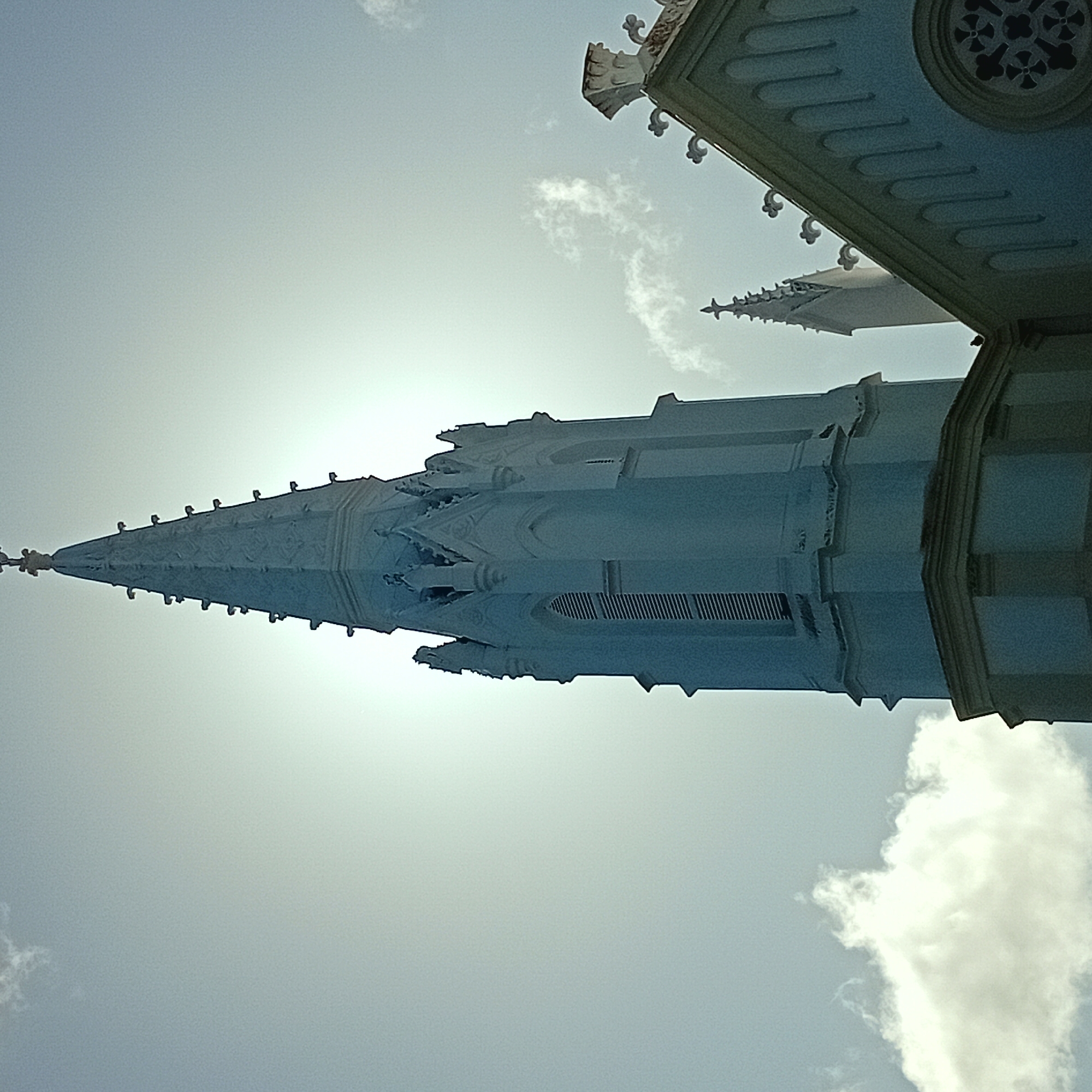 The image depicts a building's steeple, viewed from a low angle against a bright sky. The steeple is light blue-grey and features pointed spires and decorative elements. Some of the building's roof is visible at the top right, with ornate detailing. A bright light source obscures the center of the image, suggesting the sun is directly behind the structure. Clouds are present in the lower right corner of the frame. The location is not determinable from the image content.