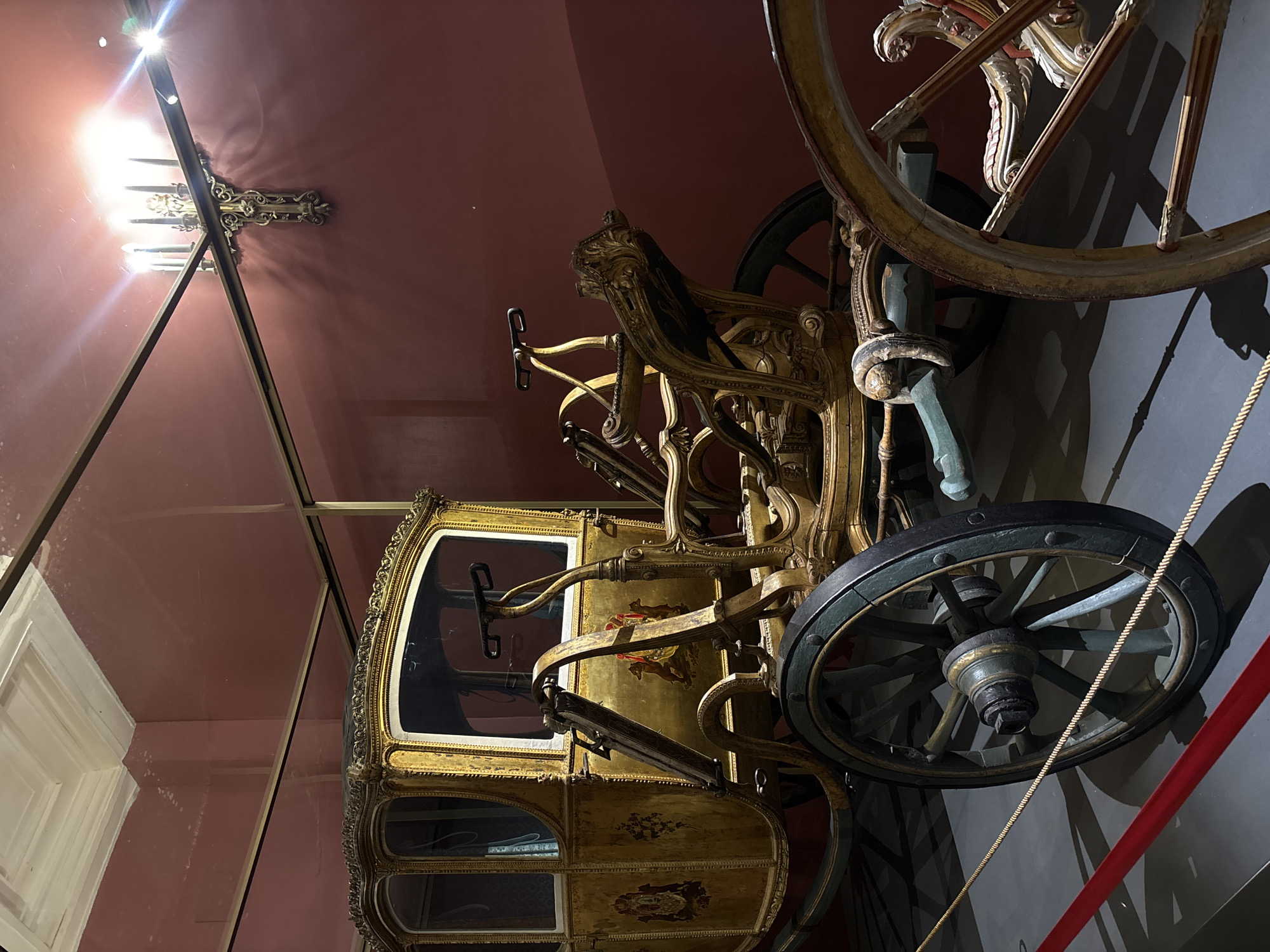The image depicts an ornate, historical carriage encased in glass. The carriage is painted in gold and other colors, featuring intricate designs and detailing. It has large wheels and a cabin with windows. The carriage is displayed inside a room with a red ceiling and grayish walls. A chandelier can be seen through the glass case. A red rope is placed near the wall. The photo was taken at the Moscow Kremlin Armoury Chamber in Moscow, Russia.