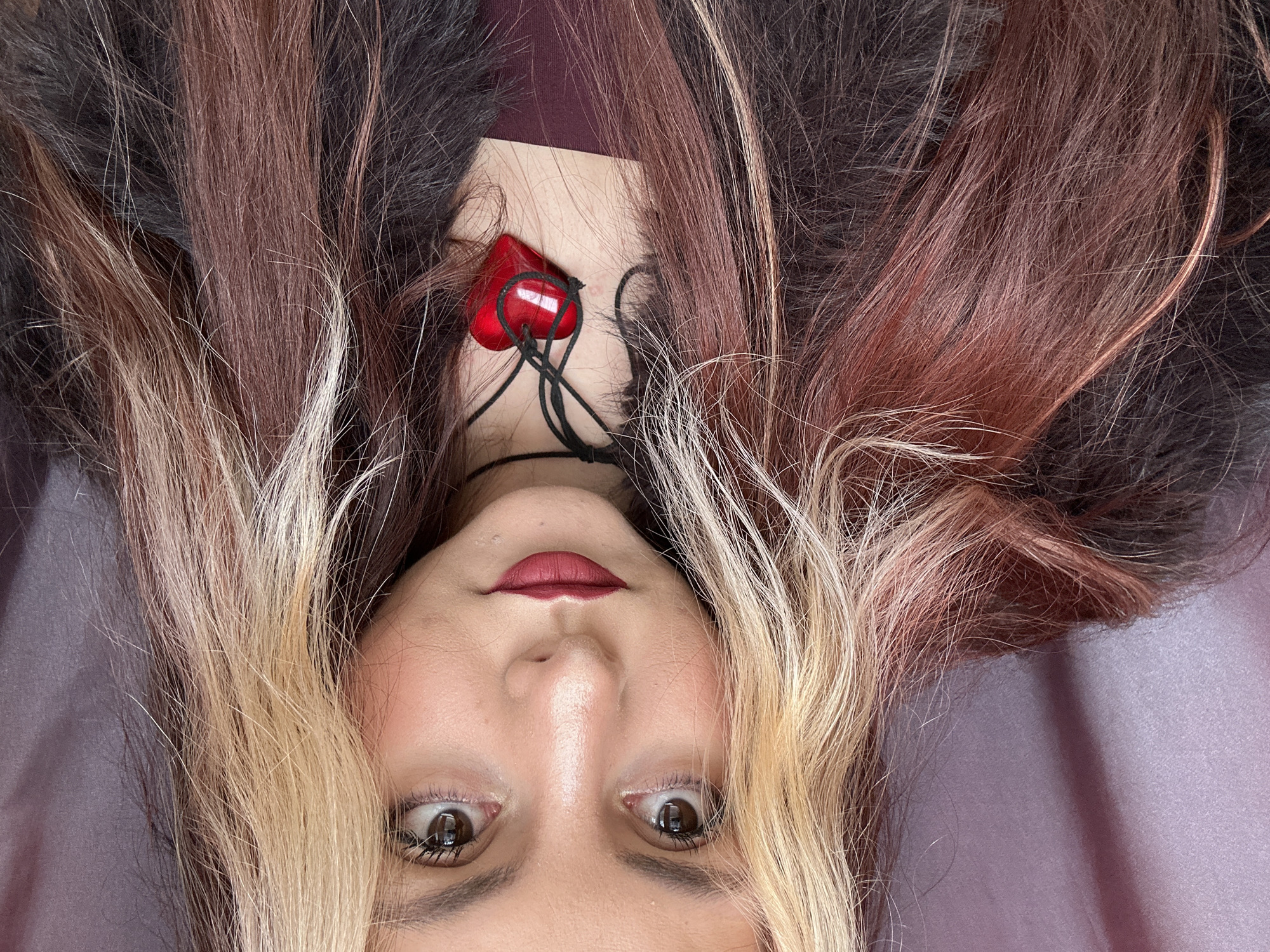 A close-up, upside-down photograph captures a woman's face, surrounded by multi-colored hair. The hair is predominantly dark brown and black, with streaks of blonde and reddish-brown. A red heart-shaped pendant hangs around her neck. The woman has fair skin, brown eyes, and her lips are painted a dark red. The background is a smooth, light purple fabric. The location is not discernible from the visual data provided.