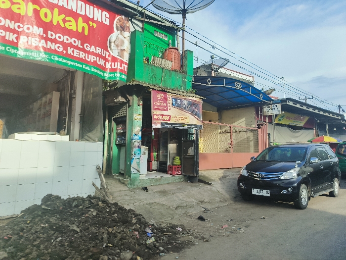 The image shows a street scene in Cikalong, Indonesia, located at the coordinates -6.735 latitude and 107.438 longitude. The scene features a small black car parked on the right side of the road, facing the camera. The car is a Toyota Avanza, identified by its license plate "D 1337 KK." The car is parked in front of a row of shops on the left side of the road. The shops are a mix of small businesses and residences. The buildings are brightly painted with green and red colors. The shops are closed for business. The scene is quiet and there are no people visible. There is a pile of dirt and debris in the foreground. The sky is partly cloudy. The environment is urban.