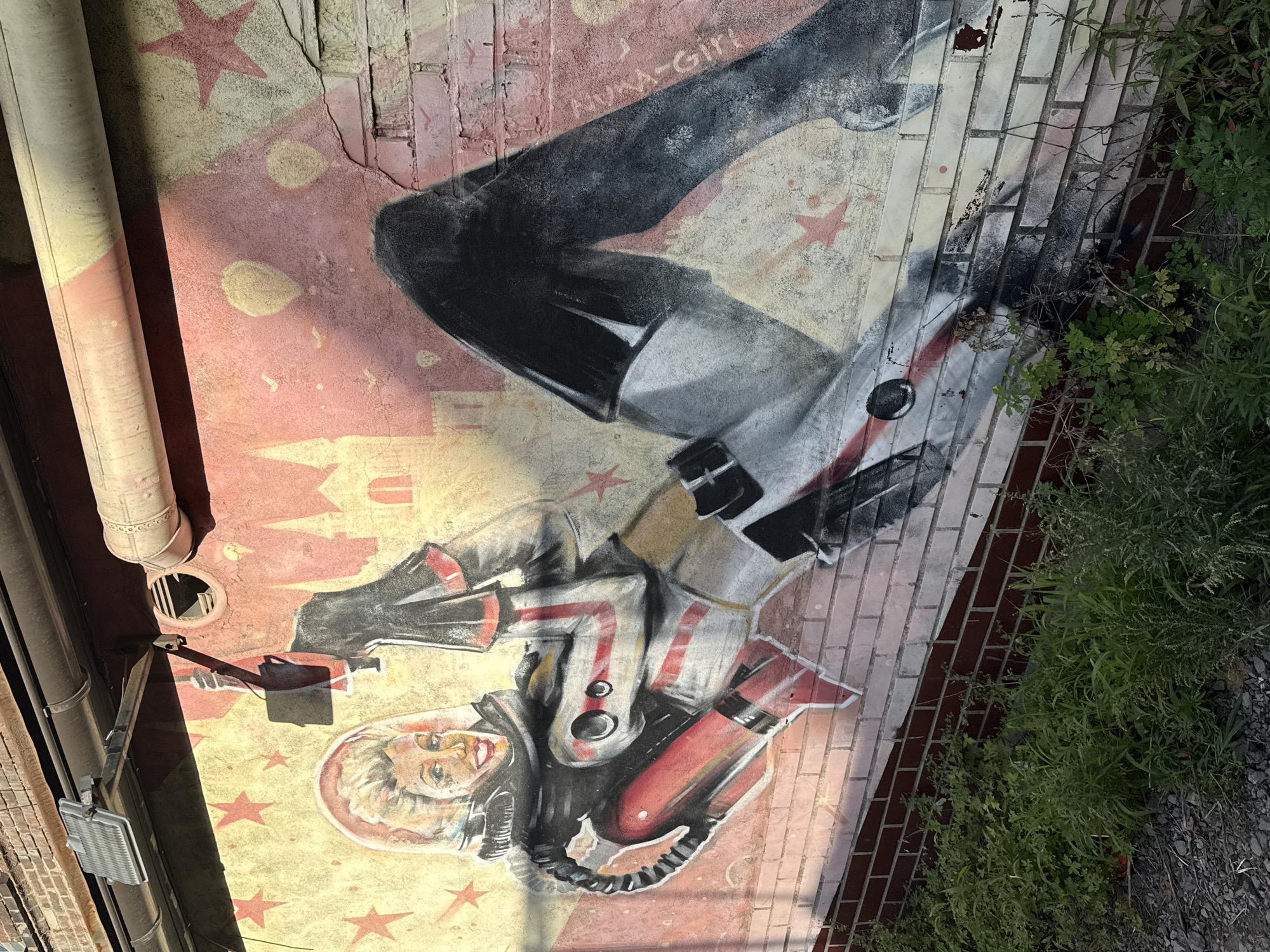 The image depicts a mural painted on a brick wall in Vladivostok, Russia. The mural features a stylized depiction of a woman in a retro-futuristic astronaut suit, lounging provocatively. She has blonde hair styled in a voluminous manner, a beaming smile, and is wearing a clear helmet. Her suit is primarily white with red and black accents, including a prominent red oxygen tank attached to her back. The background of the mural is a blend of yellow and pink, with faded red stars and silhouettes of buildings resembling castles or cathedrals. There is also a hint of what appears to be a signature or tag on the right side of the mural, reading "MOM-GIRL". The lower portion of the wall is adorned with a textured brick pattern, and the area in front of the wall is overgrown with green vegetation and gravel. Overhead, there's a dark pipe and a light fixture attached to a brick structure, casting shadows on the mural. The overall impression is one of vintage sci-fi aesthetics combined with urban decay.