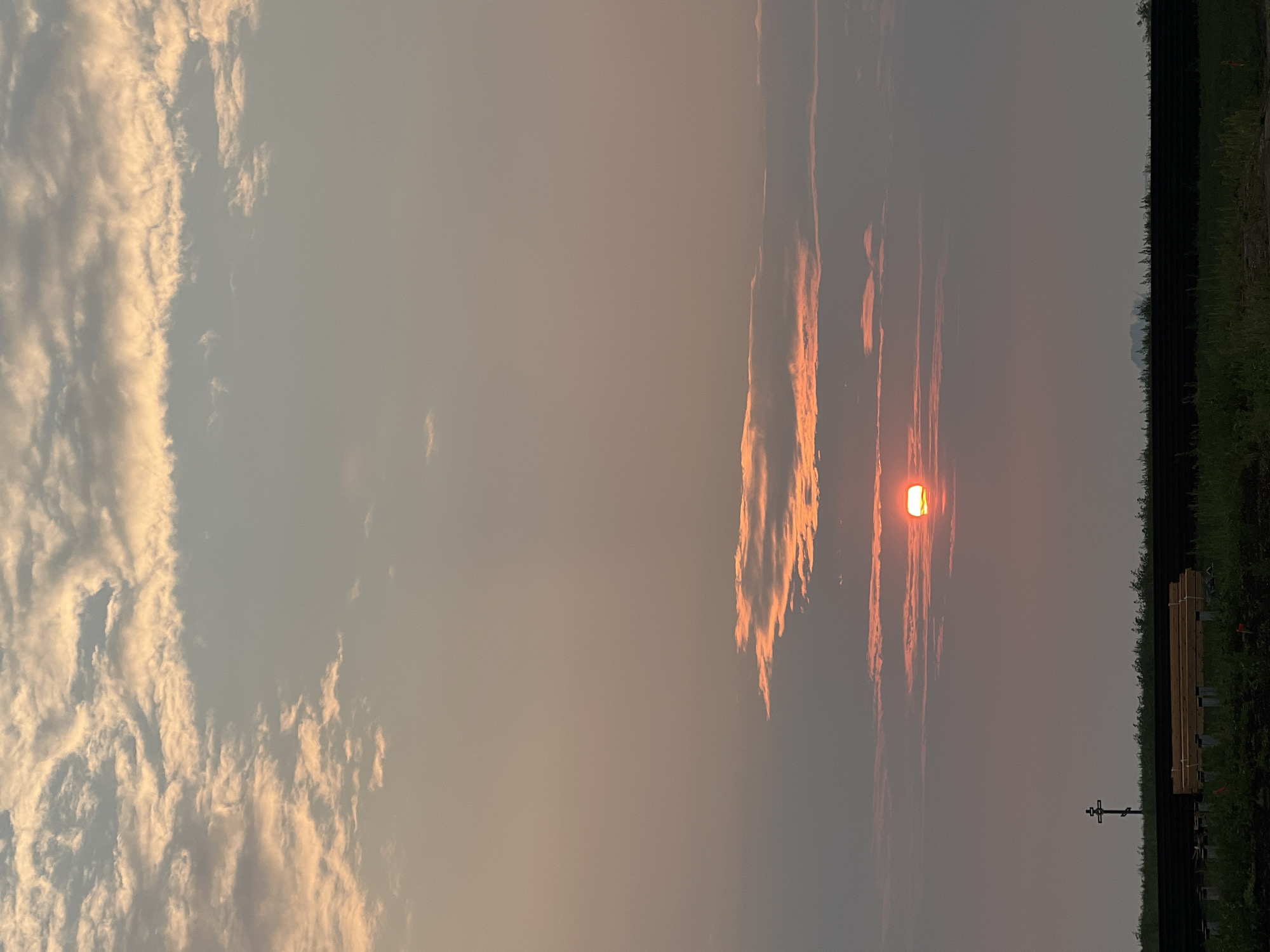 The image captures a sunset over a rural landscape, likely in the vicinity of Novopichugovo, Russia. The sky is filled with clouds illuminated by the setting sun, which appears as a bright orange disk partially obscured by wispy clouds in the middle ground. The upper portion of the sky transitions from a pale blue to a soft orange, with streaks of white and gray clouds. The lower part of the image shows a dark horizontal fence line, possibly marking a boundary. In front of the fence, there is a pile of wooden beams, suggesting construction or storage. To the left of the wooden beams, a dark, tall, cross-shaped structure is visible against the horizon. The foreground consists of green grass and some scattered vegetation. The overall atmosphere is serene and natural, with the sunset casting a warm glow over the scene.