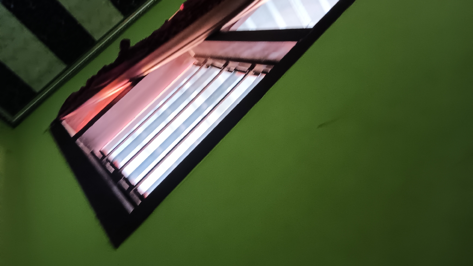 A close-up, low-angle shot depicts a green wall with a window. The window has horizontal bars and is partially covered by a pinkish curtain. The scene is dimly lit, with light filtering through the window. The upper right portion of the image shows a black and white checkered pattern on the ceiling. The overall environment appears to be indoors.