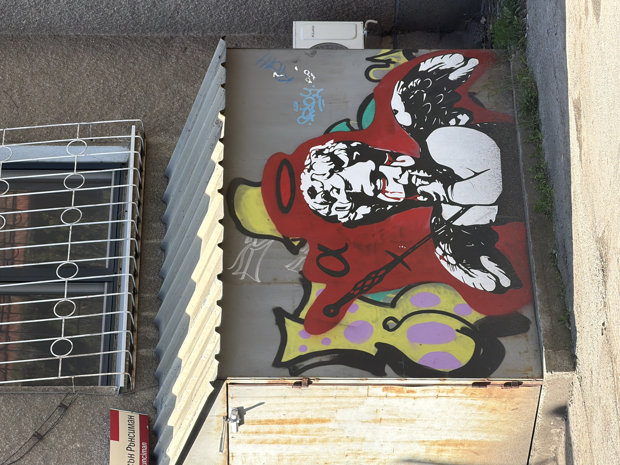 An eye-level, high-angle outdoor shot depicts a concrete wall with a metal garage door covered in graffiti. The garage is on the left side of the image and its top is covered in a layer of rusted metal. Next to the garage door is a window with a white metal grate. Above and to the right of the window, a metal garage door painted gray displays graffiti. The graffiti features a face in white and black with red outlines. The face has wings. The bottom of the face is yellow with purple polka dots and a sword through the face. Next to the graffiti is a corrugated metal overhang. Above the graffiti on the gray door is an air conditioner unit. The graffiti is located in Sofia, Bulgaria, according to text on the sign to the left of the window. Small blades of grass peek through the crack between the gray door and the concrete wall.