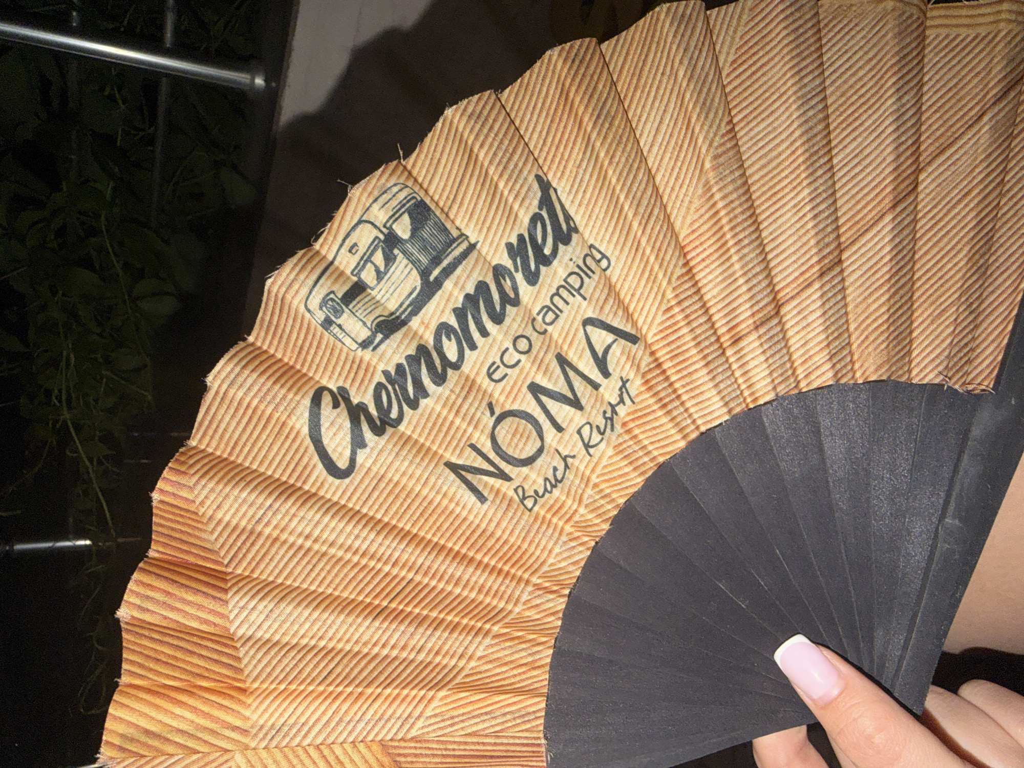 An overhead shot captures a person's hand holding a partially open folding fan in an outdoor, nighttime setting. The fan's main fabric is a light brown or beige, featuring a subtle, repeating diagonal stripe pattern. Its visible frame on the right side is constructed from dark, possibly black, ribs. A hand with a light pink French-tipped fingernail holds these ribs.
The fan's fabric displays black text and an illustration. An outline drawing of a vintage camper van is positioned above the word "Chernomorets," written in a flowing, cursive-like font. Below this, in a smaller, sans-serif font, is "eco camping." The prominent word "NÓMA" appears below, in large, bolded sans-serif letters, with "Beach Resort" printed beneath it in a smaller, cursive, and slightly italicized script.
The background is dark, indicating evening or night. To the left, blurred dark green foliage is visible. A silver metal bar, possibly part of a railing or outdoor structure, stretches horizontally across the upper left. The branding on the fan suggests a connection to an eco-camping or beach resort location.