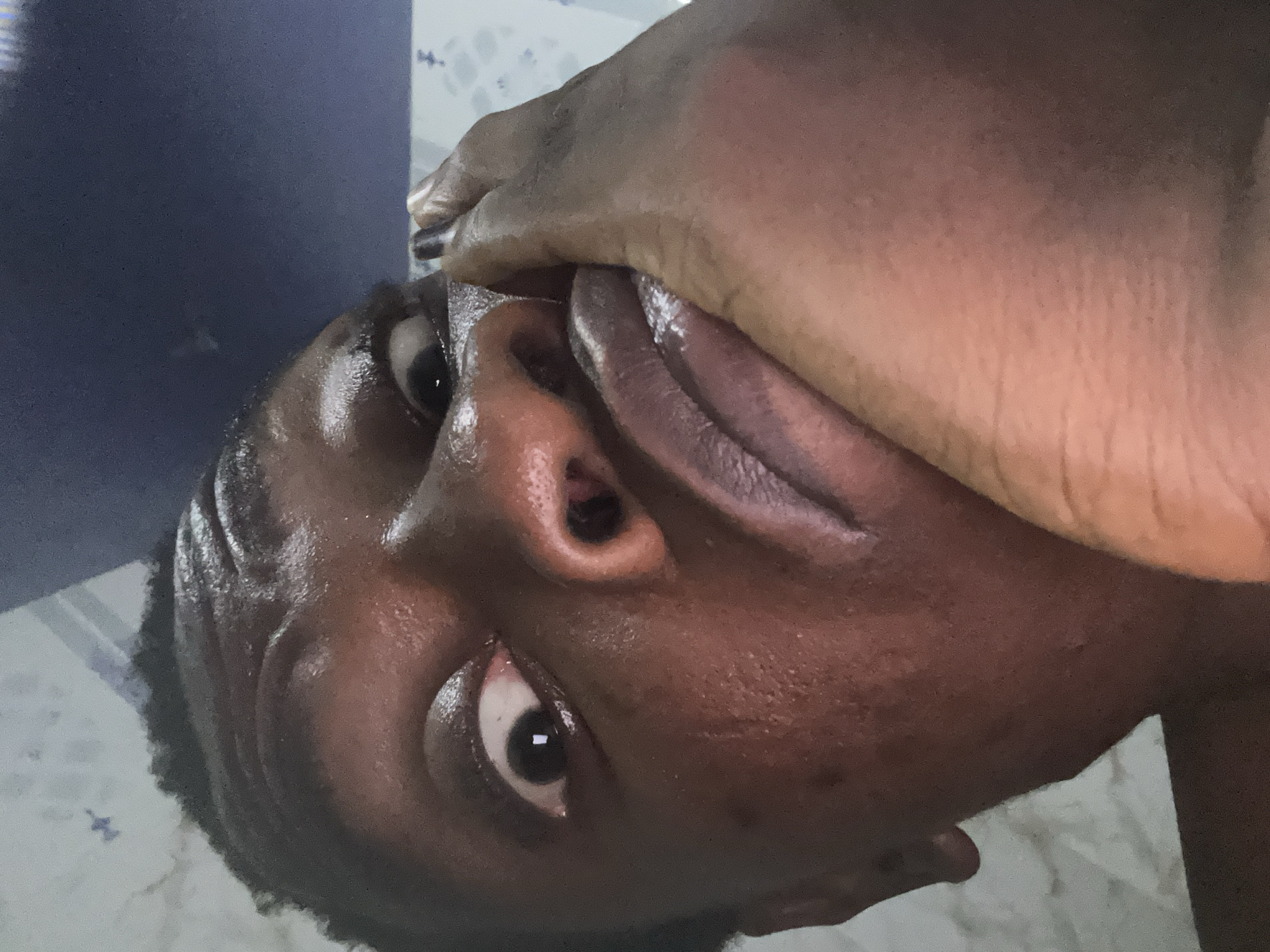 A close-up, angled, and vertically flipped shot features a dark-skinned person. Their face is shiny and wet, and their right hand is positioned over their mouth. The background shows a glimpse of a light-colored patterned surface and a dark blue, possibly textured, object. The location details are unknown.
