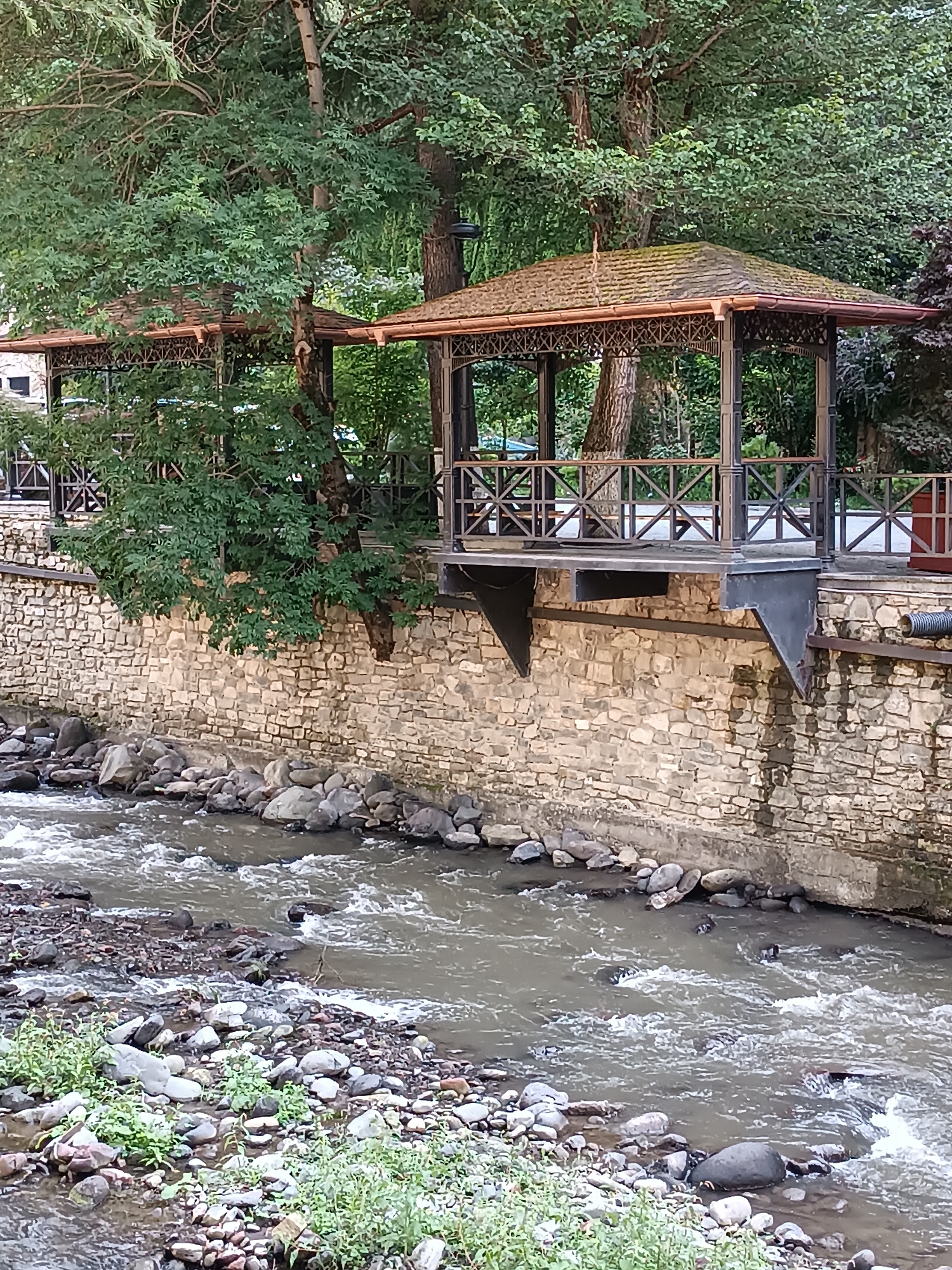 The image depicts a picturesque scene along the Borjomi River in Borjomi, Georgia. In the foreground, the shallow, rocky river flows with visible ripples and small waves. The riverbanks are lined with smooth, grey stones and patches of green vegetation.
Rising from the riverbank is a sturdy, stone retaining wall, constructed from irregularly shaped, light-colored stones. Elevated above the wall are two open-air gazebos with ornate metal framing and wooden slatted roofs. These structures are supported by dark metal pillars and beams, and feature decorative railing. Mature trees with dense green foliage partially obscure the gazebos and the surrounding environment. The overall impression is one of a tranquil, natural setting integrated with man-made structures, likely a park or recreational area. No people or animals are visible in the image.