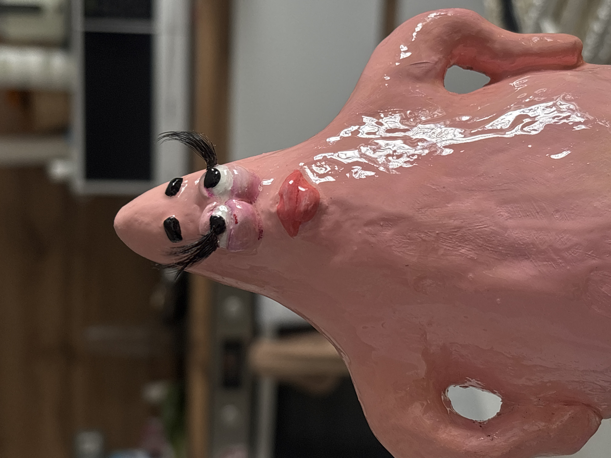 A close-up shot captures a bizarre, pink, glossy sculpture. The sculpture's head features two bulging eyes, each adorned with exaggerated eyelashes. Below the eyes, black dots are scattered across the surface. A bright red, blob-like shape represents the mouth. The sculpture's overall form is vaguely amorphous with a cutout hole near the bottom and a loop-shaped appendage at the top. The sculpture's glossy finish reflects light. The background is blurry and indistinct but shows what appears to be household appliances and furniture. There is no information or indication about the location being Krasnoyarsk, Russia.