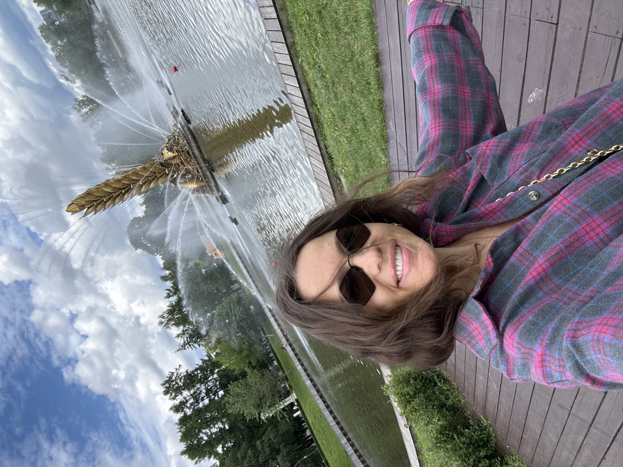 A woman wearing sunglasses and a pink and gray plaid shirt takes a selfie with a large golden fountain in the background. The fountain, which resembles a golden ear of wheat, is spraying water in a pond surrounded by trees. The woman is standing on a wooden boardwalk with grass and bushes on the left. The sky is blue with white clouds. This scene is located in Friendship of Peoples Fountain, VDNKh, Moscow, Russia.