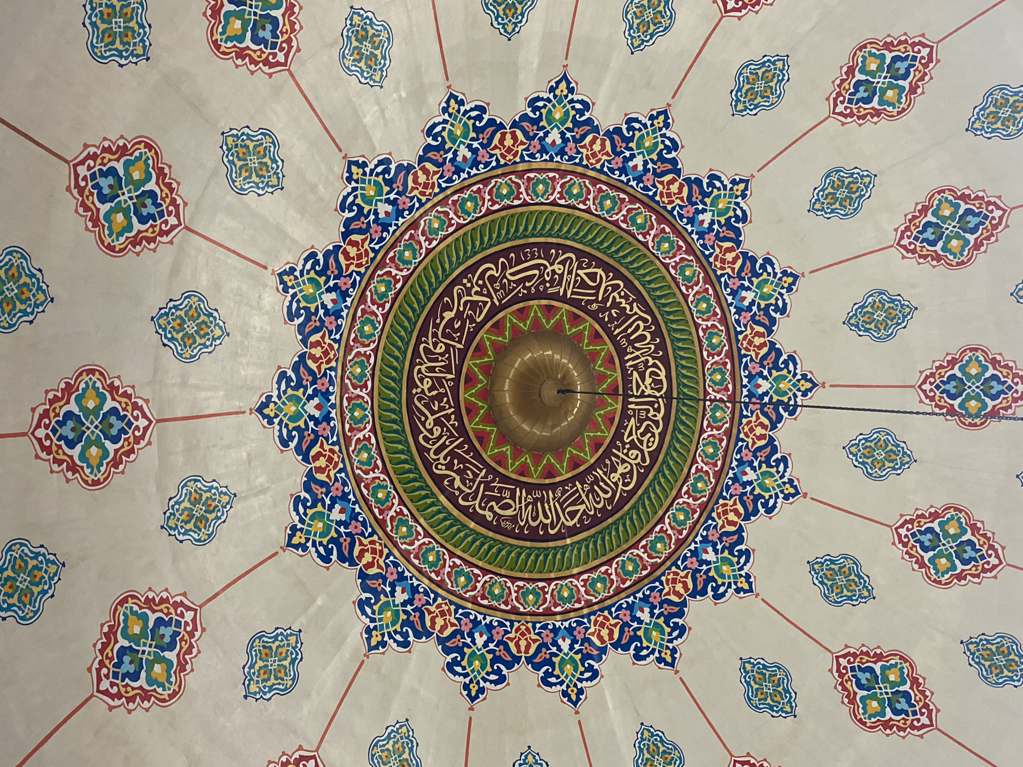 The image shows a close-up view of a highly decorated ceiling, likely within a mosque. The dominant feature is a large, intricate circular design in the center, composed of concentric rings of geometric patterns and calligraphy in vibrant colors, including red, blue, green, and gold. This central medallion appears to be an artistic representation of Arabic script. Surrounding the central design are radiating lines and smaller, repeating floral or geometric motifs in similar colors, spread across a light-colored background. A golden dome-shaped fixture, likely a lamp or part of the building's architecture, is visible in the very center of the circular design. The overall impression is one of rich Islamic art and architecture.
While this image displays elements commonly found in mosques, its specific location in Lhokseumawe, Indonesia, cannot be definitively determined from the visual information alone. This type of artwork is prevalent in many mosques across the Islamic world.