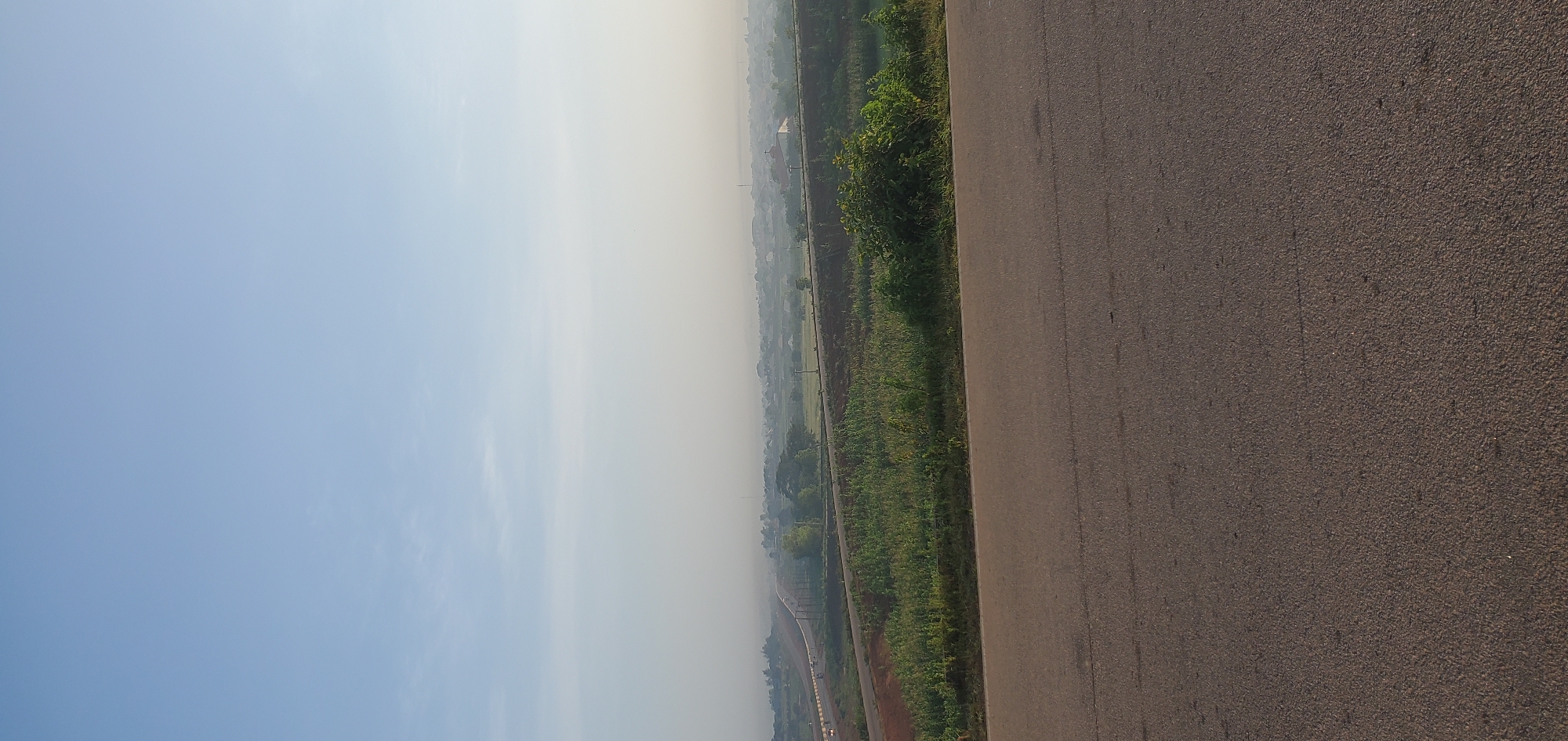 This is a vertical, medium-shot photograph of a hazy landscape in Kakau, Nigeria, taken from an elevated position looking towards the horizon. In the foreground, a dark asphalt road stretches across the bottom of the frame. Beyond the road, a vibrant green embankment is covered with dense vegetation and scattered bushes. Further into the distance, a multi-lane highway is visible, with some vehicles appearing as small specks. The landscape transitions into a hazy expanse of small buildings, scattered trees, and open fields, suggesting a rural or semi-urban setting. The sky above is a pale blue with a slight haziness, indicating either early morning or late afternoon light. No people are discernible in the image.