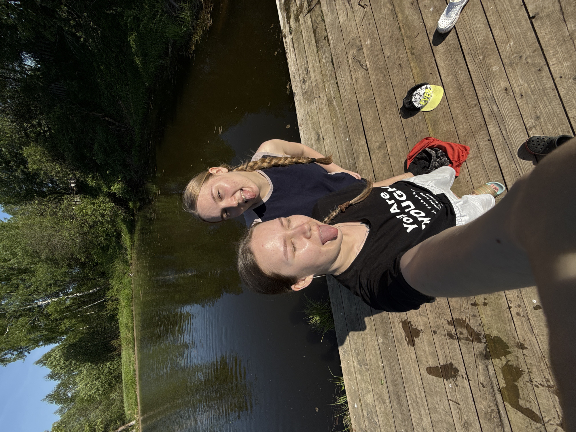 Two women are taking a selfie on a wooden deck near a body of water. One woman wears a navy tank top, while the other wears a black t-shirt with white text. Both women have their tongues sticking out at the camera. A body of water is on the left, surrounded by green trees. On the wooden deck, there are shoes, a cap, and a red piece of clothing. There are puddles of a liquid on the deck. The location is in Zhigalki, Russia.