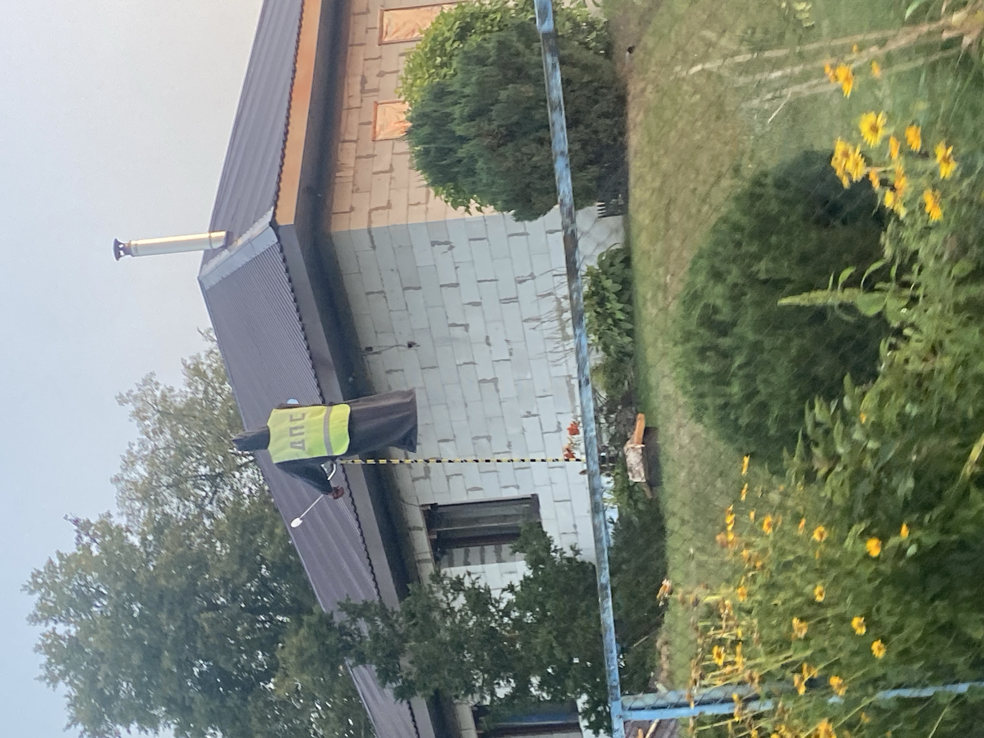 The image captures a scene in the village of Vovchyn, Belarus, featuring a house with a metal roof and cinder block walls. A mannequin, dressed in a dark hooded robe and a bright yellow vest with "ДПС" written on it in Cyrillic, is positioned on the roof. The mannequin holds a white rod with a spray nozzle. A black and yellow striped cord hangs vertically from the mannequin. In the foreground, a blue metal fence runs horizontally across the frame, with yellow flowers and green foliage visible beyond it. Trees and bushes surround the house, and the sky is overcast.