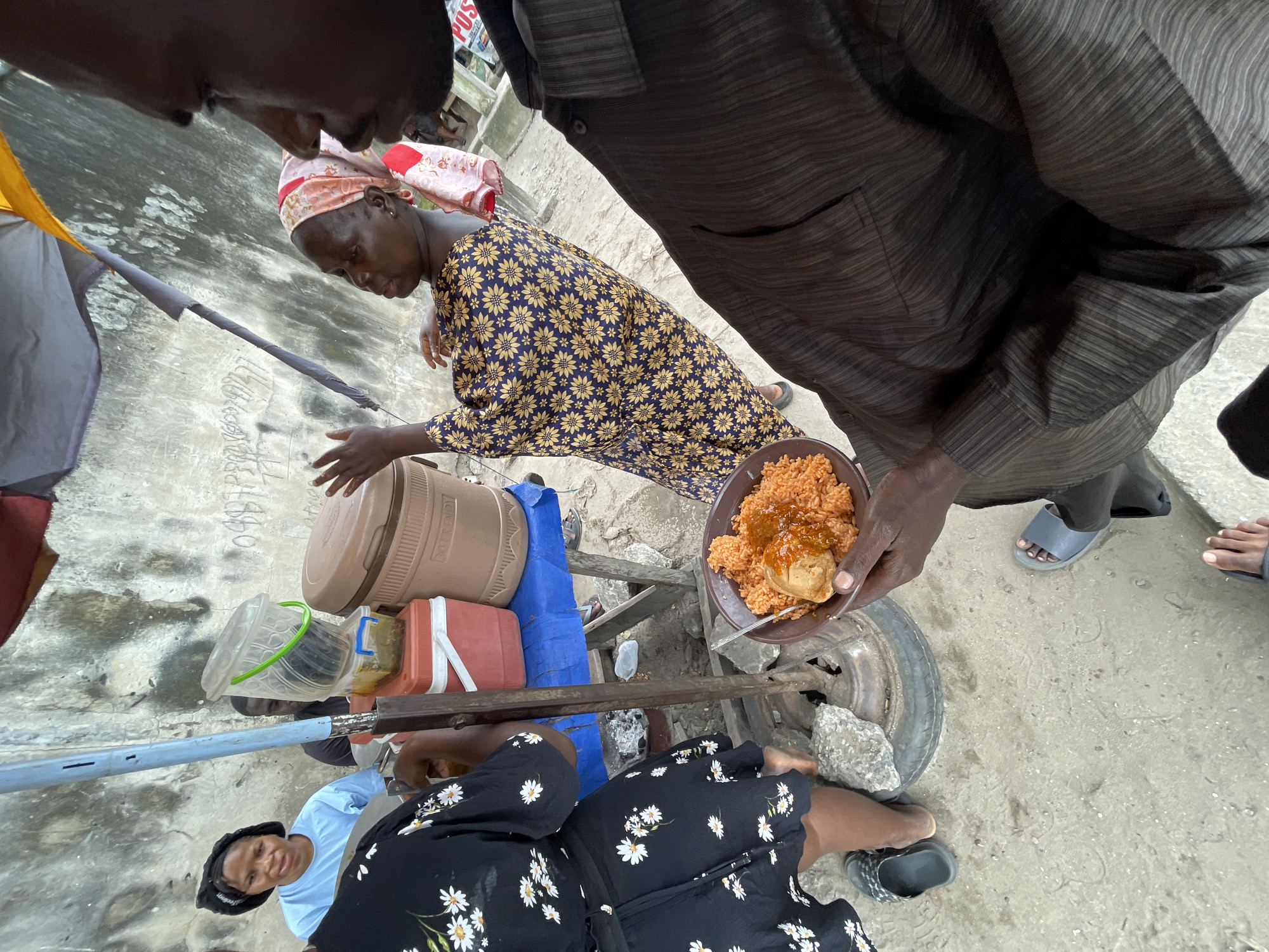 The image depicts a street food vendor scene in Ado, Nigeria. In the foreground, a person's hands hold a bowl of jollof rice with a piece of fried chicken on top, viewed from a first-person perspective. The background reveals a food stall with various containers, including a large brown insulated cooler, red and white coolers, and clear plastic tubs, one with a green handle. A woman in a blue t-shirt and black headwrap is seated behind the stall. Another woman, wearing a yellow and blue patterned dress and a pink and white headscarf, appears to be interacting with a customer, gesturing with her hands. A man in a dark striped shirt stands close to the food stall, partially obscuring the view. The environment is characterized by a rough, textured wall in the background and a concrete ground surface. The overall scene suggests a busy commercial area on a street.