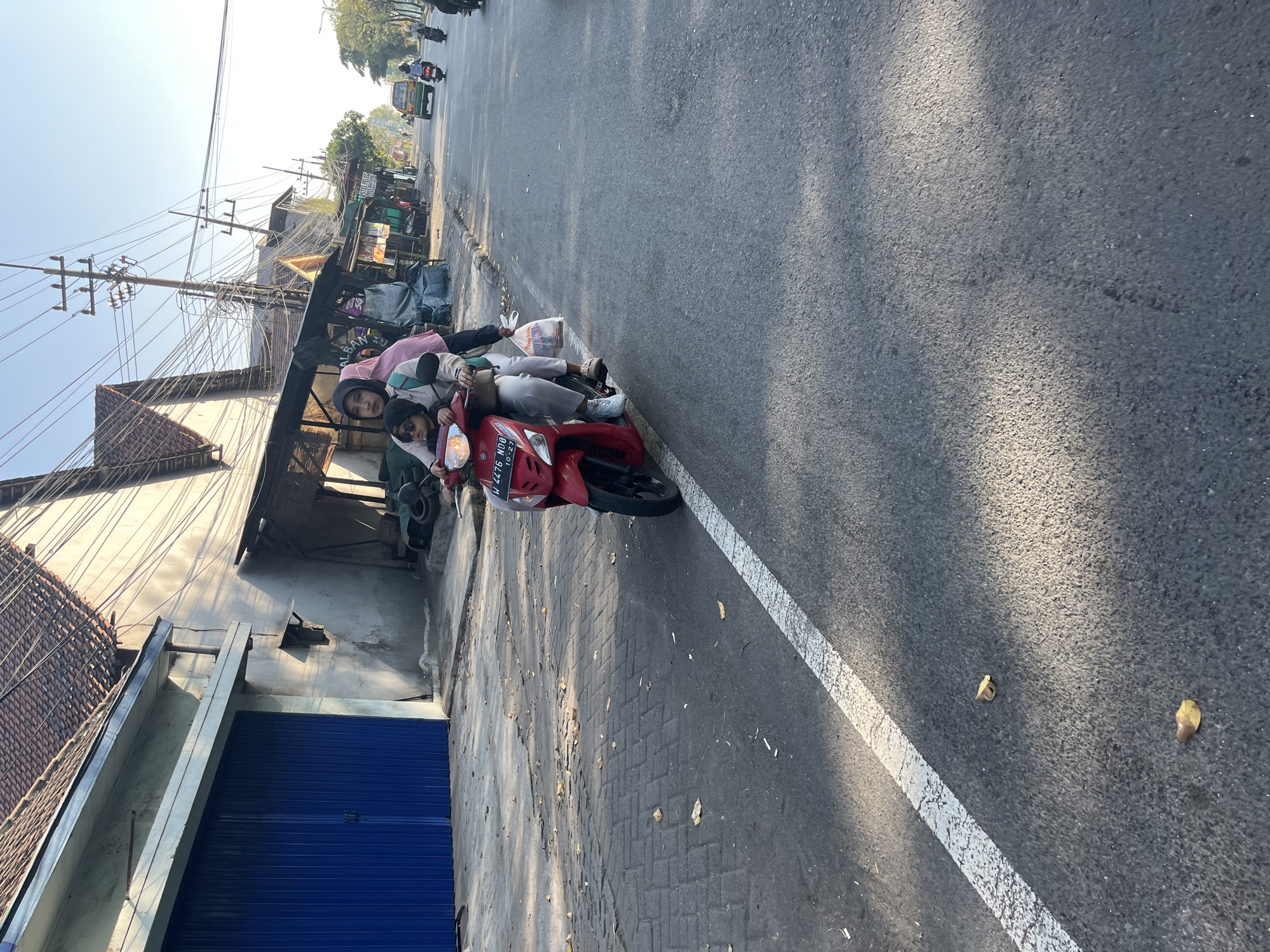 In Kludan, Indonesia, on a sunny day, three individuals are on a red motorbike traveling on a paved road. The driver, a woman wearing a light pink headscarf, is in the front. Beside her, a child in a dark beanie and sunglasses is seated. Behind them, another woman, partially visible, holds a plastic bag. The motorbike's license plate reads "W 4476 NDO 10-23". The surrounding environment includes buildings with tiled roofs, shops with awnings, and utility poles with numerous wires. The road has a white line marking its edge. Leaves are scattered on the sidewalk and road. In the background, other vehicles, including a green car and more motorbikes, are seen on the road, indicating a bustling urban setting.