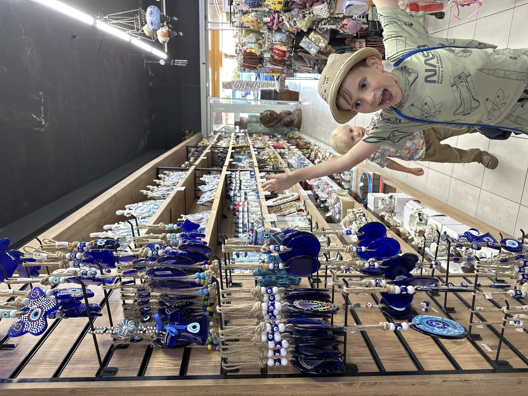 A high-angle indoor shot depicts two young boys in a shop in Fethiye, Turkey. The older boy, positioned closer to the camera on the right, wears a straw hat and a green long-sleeved NASA t-shirt. He is smiling broadly, and reaching out with his left hand. He has a blue string bag across his shoulder. The younger boy, slightly blurred in the background behind the older boy, has blond hair and is wearing a patterned shirt and tan trousers.
The foreground features displays of blue evil eye charms, attached to twine and arranged on a black metal grid atop wooden shelves. The items are presented in a closely packed array.
The background displays a range of merchandise on wooden shelves. A black wall is visible on the left, with lighting fixtures and chains. On the right, beyond the shelves, there are bags and other items hanging from the ceiling. The floor is tiled in light colors.