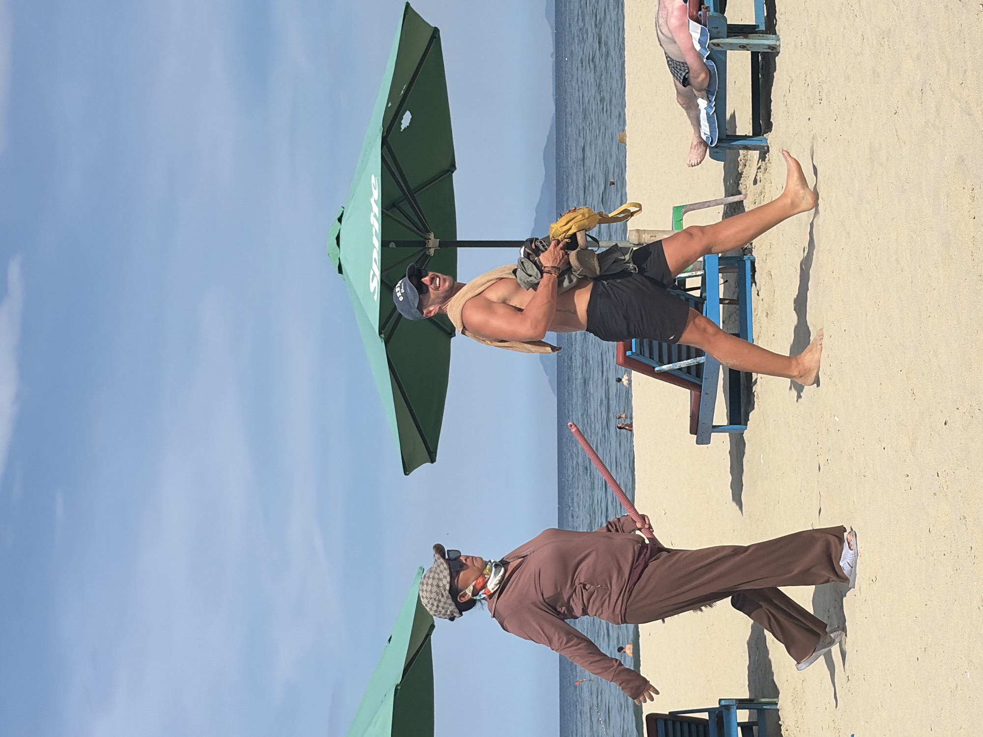 This is a full-shot, eye-level photograph taken on a sandy beach in Đà Nẵng, Vietnam, with the ocean and sky as the background. Two people are walking on the beach, and beach chairs and umbrellas are visible.
In the foreground, on the left, a person wearing a brown tracksuit, a patterned baseball cap, sunglasses, and a patterned face covering is walking to the left. They are holding a long, pink rod in their right hand.
To the right of this person, a shirtless man wearing black shorts, a blue baseball cap, sunglasses, and a towel draped over his shoulders is walking to the right. He is carrying a bundle of clothing or items in his arms.
In the background, a person is lying on a blue beach chair under a green umbrella. Another person is partially visible on a similar beach chair in the far right of the frame. The ocean stretches out behind the beach, and distant mountains are visible on the horizon. The sky is bright blue with some scattered white clouds. The green umbrellas have the "Sprite" logo visible on them.