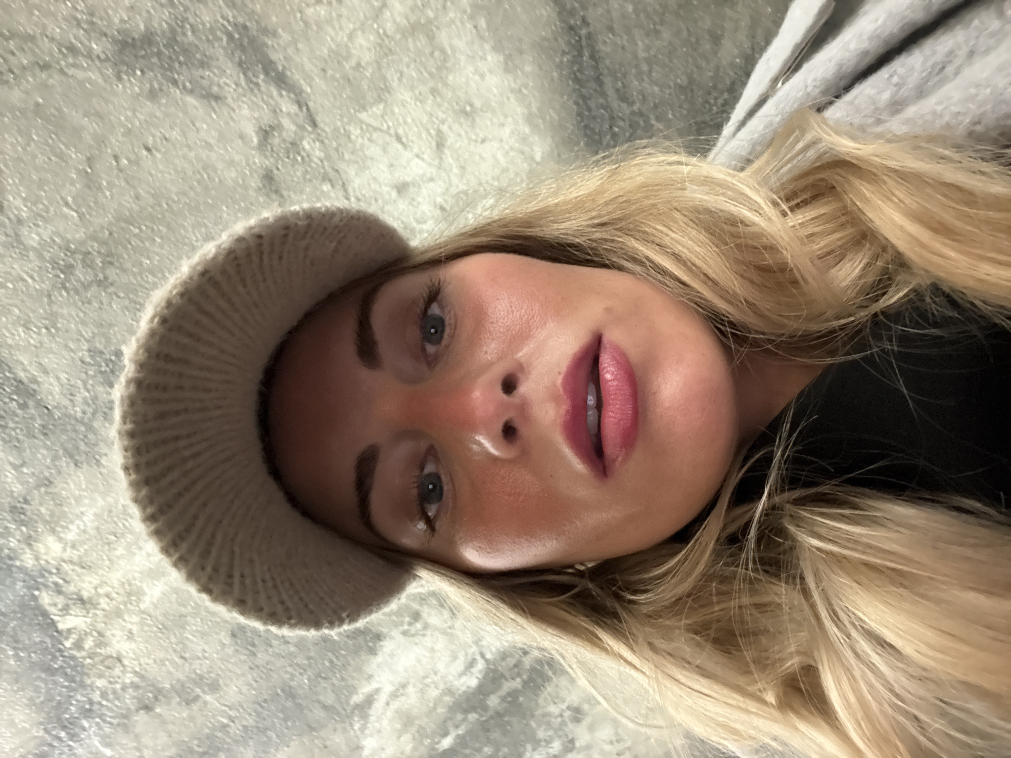 A selfie photograph shows a fair-skinned woman with blonde hair wearing a beige knitted hat. The woman's head is tilted in the frame, showing her face from below. Her eyes are blue, and her lips are painted a deep pink. The background is a textured gray surface, possibly a wall. There is no evidence to indicate the location is Paris, France. The photograph does not contain sufficient details to indicate any subjects, objects, actions, or interactions.