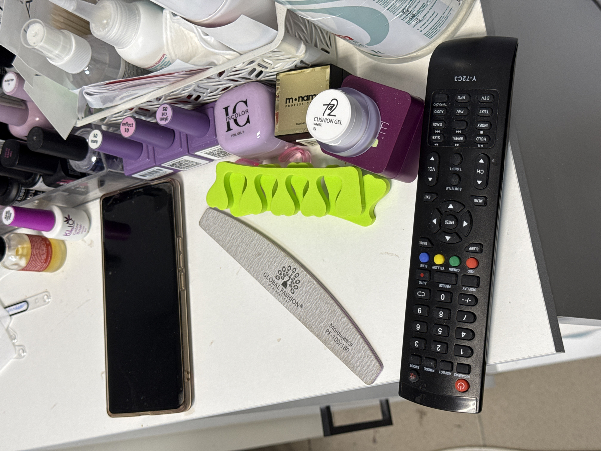 The image shows a cluttered white surface, likely a desk or shelf, with various personal care and electronic items. On the left, a black smartphone is placed horizontally. To its right, a light gray nail file with text indicating "GLOBAL FASHION®" and "MONOLIJSER PF-100/180" is positioned diagonally. A bright green, plastic toe separator lies to the right of the nail file. Further to the right, there is a collection of nail polish bottles, primarily in shades of purple and pink, some with black caps. One bottle has "KLIO" and "Color Base" visible. There is also a small white jar labeled "772 CUSHION GEL WHITE" sitting on a purple container. A gold-colored box with black text reading "m² nam" is visible behind the gel jar. In the background, a white plastic basket holds more items, including what appears to be a spray bottle and wooden sticks. Dominating the bottom of the image is a black television remote control with numerous buttons, including a power button, number pad, and various function keys. The remote has the model number "Y-72C3" printed on its lower right. A partially visible large white bottle with green and blue text is on the far right edge of the frame. The lighting suggests an indoor setting. There is no indication of the city of Емельяново, Russia, within the image itself.