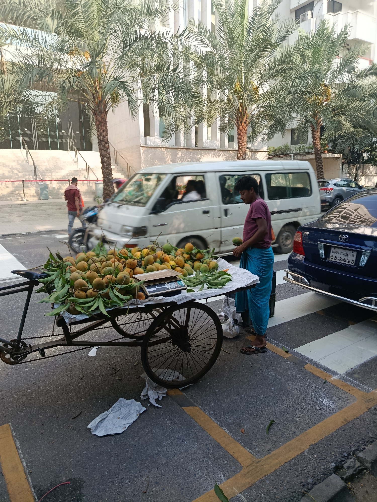 A street scene in Dhaka, Bangladesh, features a vendor selling mangoes from a three-wheeled cart. The cart, laden with mangoes and leaves, includes a scale for weighing the fruit. The vendor, wearing a maroon shirt and blue lungi, stands beside the cart holding a mango. Behind him, a dark blue sedan is parked, with its license plate visible. A white van drives past in the background, and a pedestrian is crossing the street further away. Palm trees line the sidewalk. The road surface is asphalt marked with yellow lines and pedestrian crossings. Discarded paper lies on the ground near the cart.