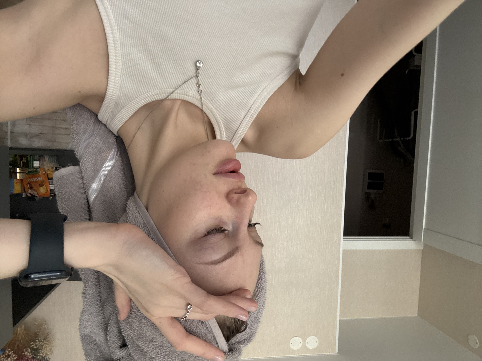 A young, light-skinned woman with a towel wrapped around her head is depicted in an upside-down photograph. She is wearing a light-colored tank top and a necklace with a pendant. Her left hand is raised to her face, with a ring visible on one of her fingers. She is wearing a smartwatch on her left wrist. The background includes a wall, a window, and a glimpse of a room with various objects. The location is potentially within an apartment or residential setting. The image contains insufficient information to pinpoint the location within Perm, Russia.
