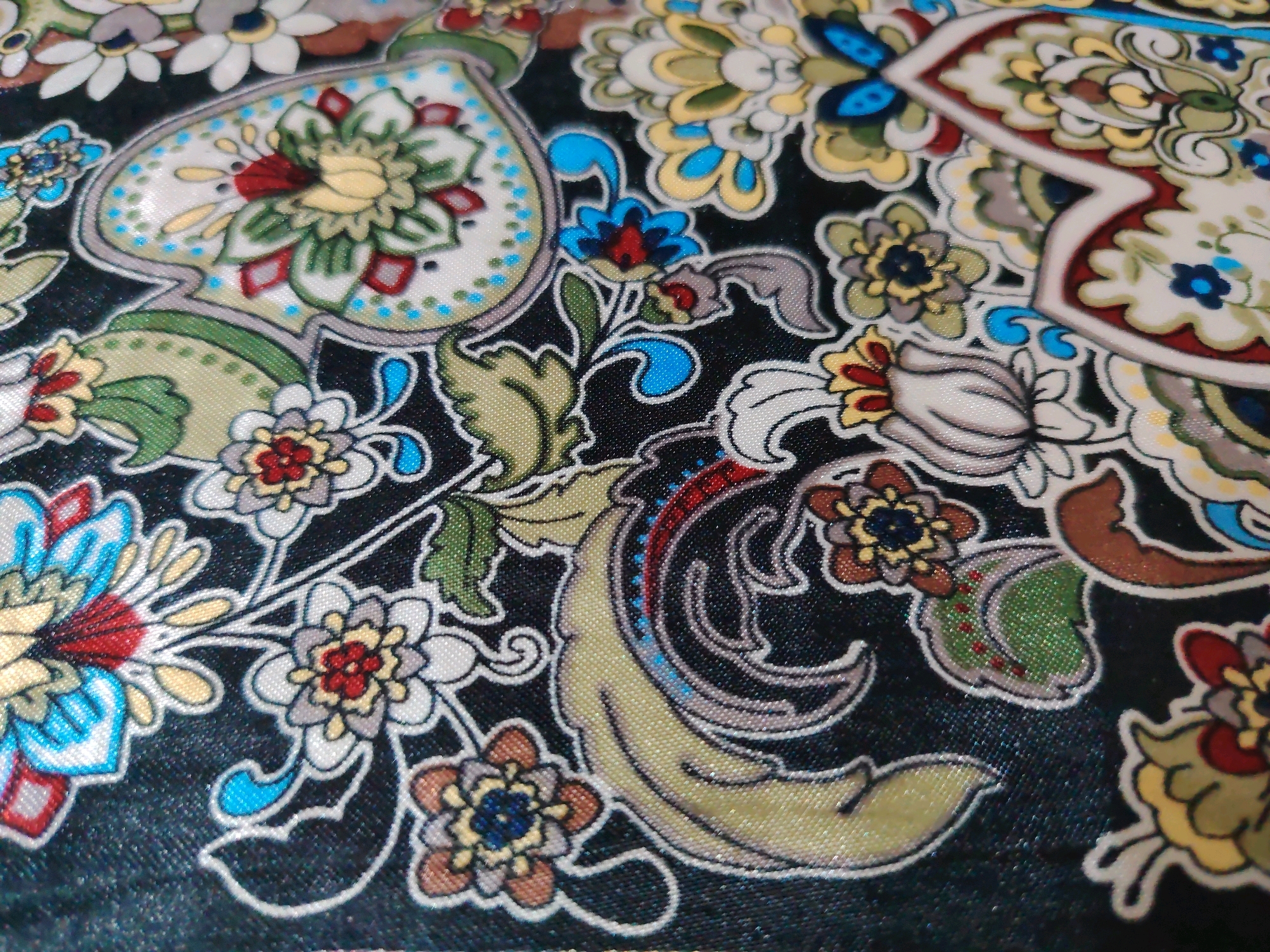 The image displays a close-up, overhead view of a patterned fabric, likely a textile or garment, with a dark, possibly black, background. The pattern is ornate and features stylized floral and paisley motifs. The colors are vibrant, including shades of yellow, green, blue, red, and white, outlined with thin black lines. The texture of the fabric appears to be slightly reflective, suggesting a synthetic material or a sheen finish. There are no people, actions, or interactions visible in the image. The scene is a flat, two-dimensional surface, with no environmental context to indicate a specific location within Davao City, Philippines or any other place.
