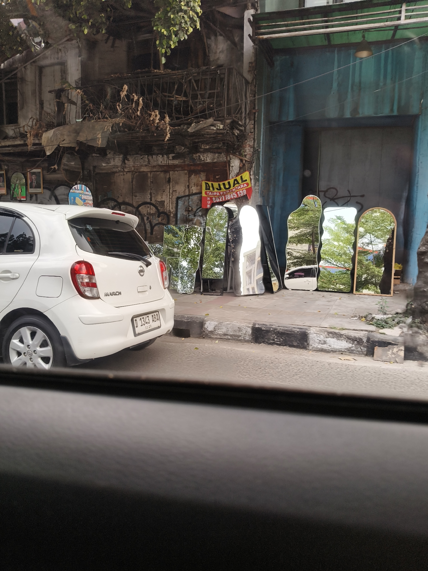A white Nissan March car is parked on the side of a street in Bandung City, Indonesia, alongside several irregular-shaped mirrors propped up against a building. The building appears aged and somewhat dilapidated, with exposed pipes and balconies. A yellow banner with Indonesian text is visible on the wall, possibly advertising a sale. Trees and greenery are reflected in the mirrors, indicating an outdoor setting. The street is paved with concrete and has a curb. The overall scene suggests a retail or display area for the mirrors on a city street.