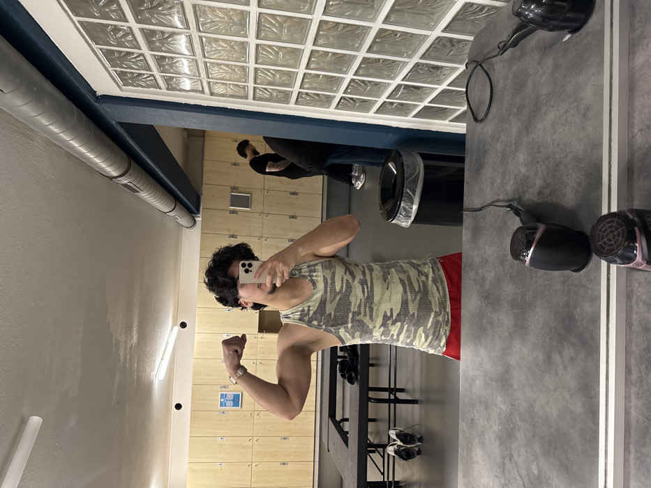 The image displays a mirror selfie captured within a locker or changing room setting, located in Geispolsheim, France. The primary subject, a male individual with dark, curly hair and light facial hair, stands centrally, reflected in the mirror. He is wearing a camouflage-patterned ribbed tank top and red shorts, and has a watch on his left wrist. He flexes his right bicep while holding a white-cased iPhone in his left hand, angled towards the mirror to take the photograph.
The background reflection reveals rows of light-colored wooden lockers. To the left, a black, rectangular bench is present, upon which dark clothing items are placed. A pair of white athletic shoes rests on the floor beneath the bench. To the right, a black trash can with a clear plastic liner is visible. Two additional individuals are present in the background: one male, dressed in a black t-shirt and dark pants, is seen looking downwards, while another figure is partially obscured by a glass block structure, with only their lower body and light-colored athletic shoes distinctly visible.
The environment features a light-colored concrete floor, a ceiling with visible fluorescent strip lighting, and exposed metallic ventilation ducts. A blue-painted structural beam runs horizontally across the upper right portion of the scene, adjacent to a wall-like structure constructed from clear glass blocks. In the immediate foreground, on the surface beneath the mirror, two black hair dryers with rose gold accents and their associated power cords are present.
