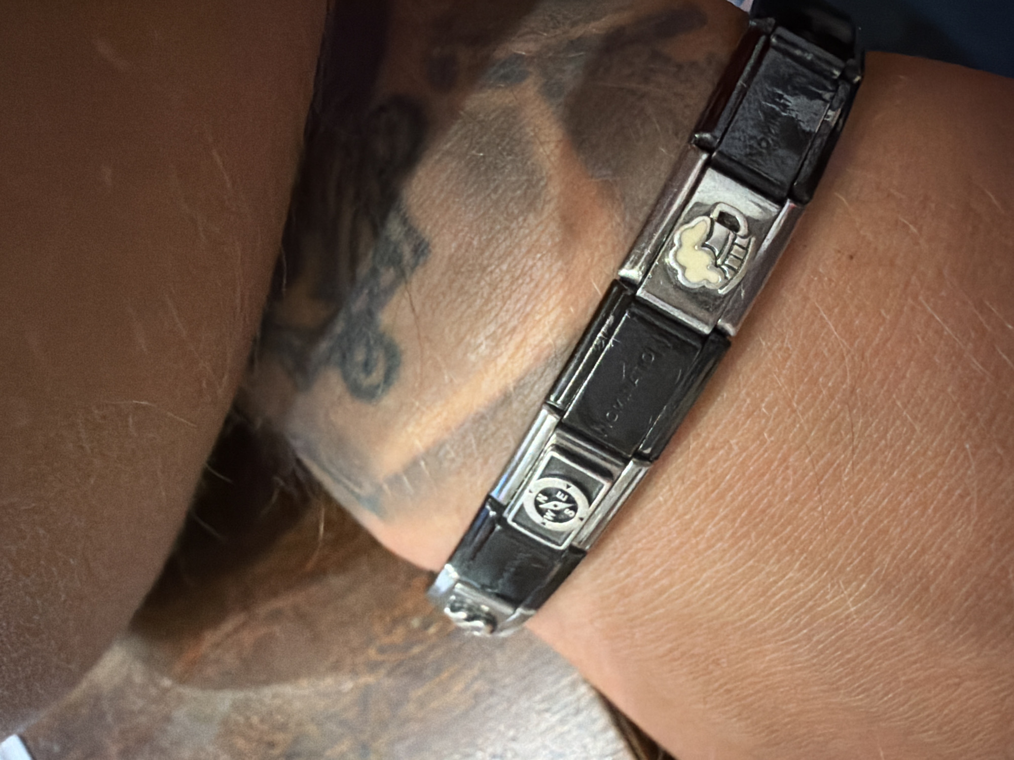 A close-up, overhead, slightly right-angled shot focuses on a person's wrist wearing a black modular charm bracelet. The bracelet features several silver-tone links with inset black enamel charms, interspersed with smaller silver-tone links. Visible charms include a compass and a beer mug. The person's skin appears to be lightly tanned and exhibits fine wrinkles and what look like light scars. A portion of a tattoo is visible on the wrist and forearm. The background is blurred and suggests a wooden surface and possibly fabric. The image is taken in an indoor setting, with natural lighting. The location is Unknown, Unknown.