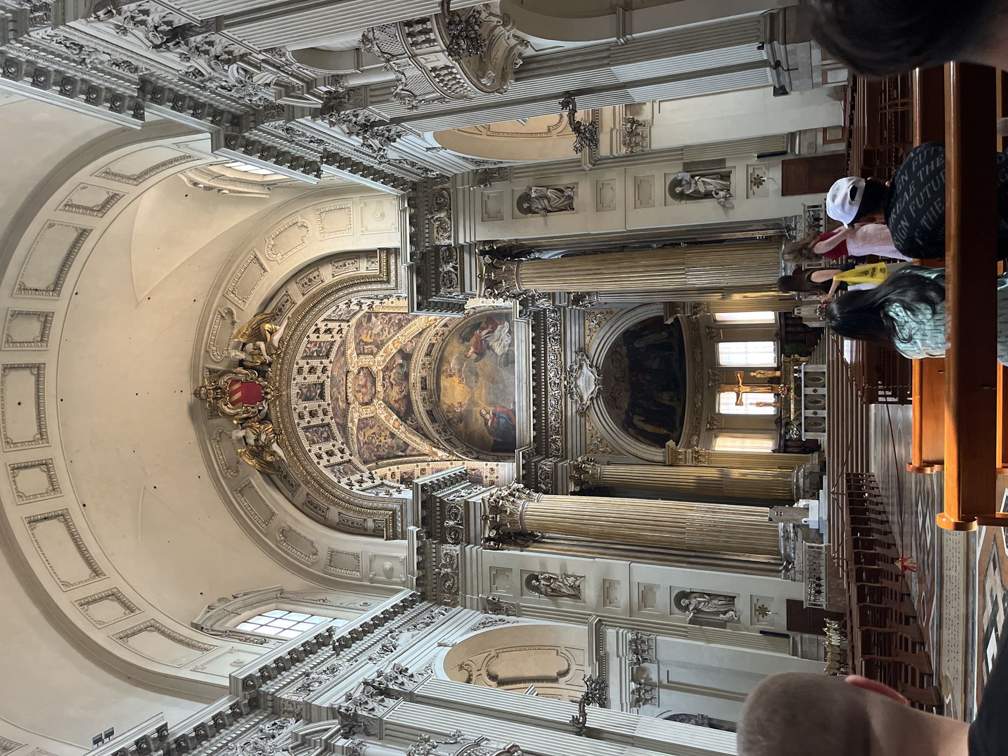 The image shows the interior of the Basilica di San Petronio in Bologna, Italy. The view is directed upwards, showcasing the basilica's ornate ceiling, complete with painted frescoes, gold detailing, and architectural embellishments. Tall, fluted columns rise to support the structure, adorned with sculpted figures. Along the side of the church, rows of wooden pews are visible, along with a few individuals who appear to be tourists or visitors observing the interior. Windows allow natural light to filter into the basilica, illuminating the scene. The building's architecture features a mix of white and gilded surfaces, with intricate designs and patterns covering the walls and ceiling.