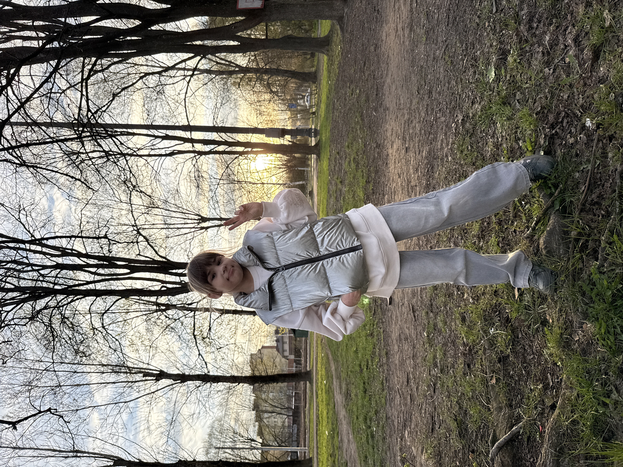 A person stands in a park setting. The individual is wearing gray pants, a white sweatshirt, and a shiny, light-colored puffer vest. They are posing with their right hand raised in a peace sign. The background shows trees without leaves, suggesting it might be late fall or early spring. Sunlight filters through the trees, creating a bright, diffused light. The ground is covered in dirt and patches of grass. Buildings are visible in the distance. Although it cannot be definitively confirmed from the image, the park's characteristics are consistent with a location in Rusanovo, Russia, based on typical urban park layouts and architecture.