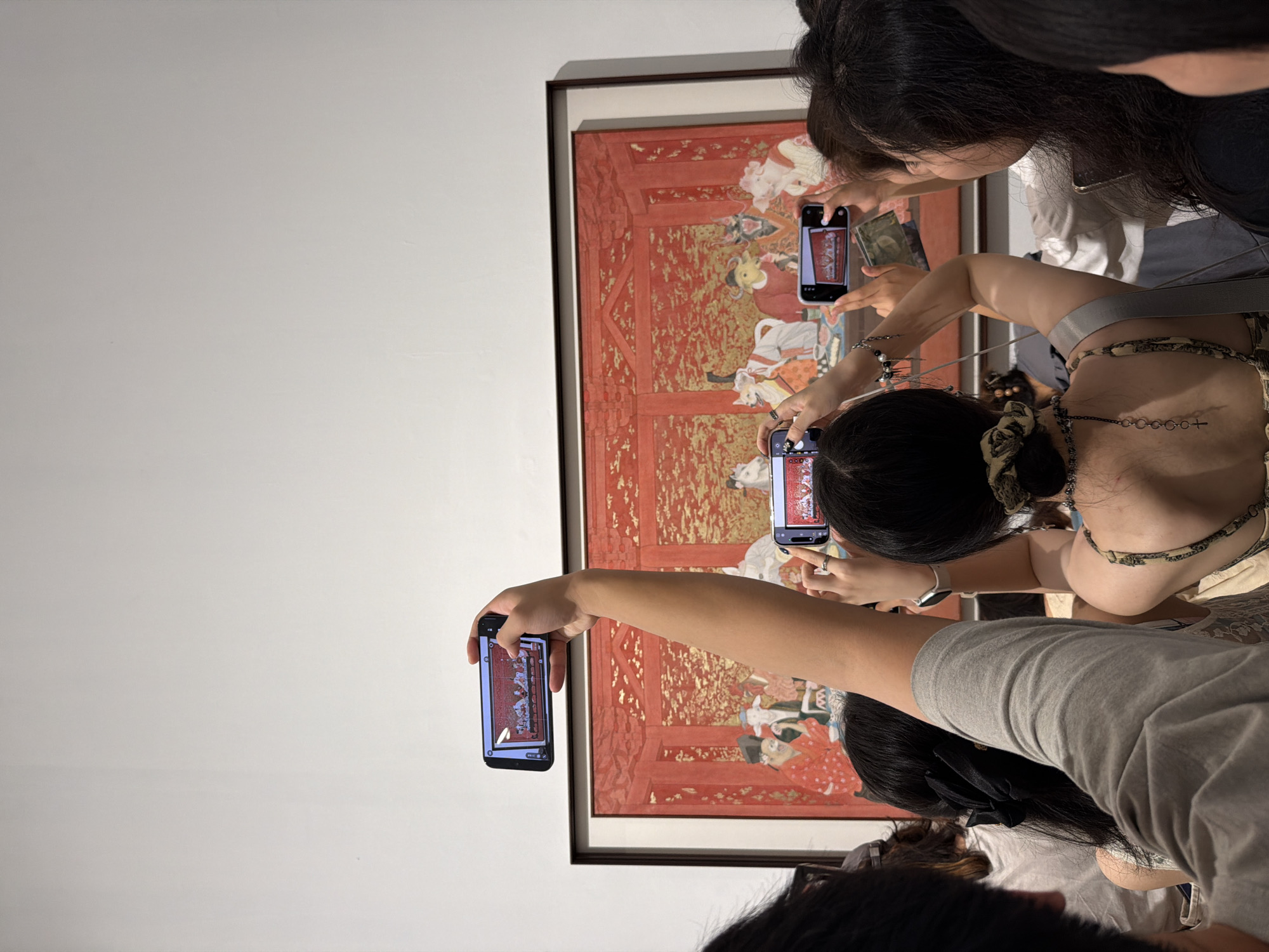 This image depicts an indoor art exhibition scene in Chongqing High-tech Zone, China. A large, rectangular painting, framed in a dark material, is prominently displayed on a plain white wall. The artwork features a vibrant red and gold color palette, portraying what appear to be several anthropomorphic animal-like figures, including a pig, an ox, and a dog, alongside human figures in traditional attire, seated around a table in an elaborately decorated room.
Multiple individuals are present, observed mostly from behind, actively engaging with the painting. At least four smartphones are held aloft horizontally by various viewers, their screens clearly displaying a real-time capture or photograph of the artwork. One person, positioned centrally in the foreground, wears a grey t-shirt and extends their arm upward to hold a smartphone directly in front of the painting. Another individual, partially visible on the right, wears a patterned strap top and a watch on their left wrist, using both hands to steady their phone. Other viewers' heads and shoulders are visible, suggesting a crowded viewing experience, with discernible hair accessories such as a black bow and an olive-patterned scrunchie. The overall environment is well-lit, with the stark white wall serving as an unadorned backdrop to the framed artwork and the interacting spectators.