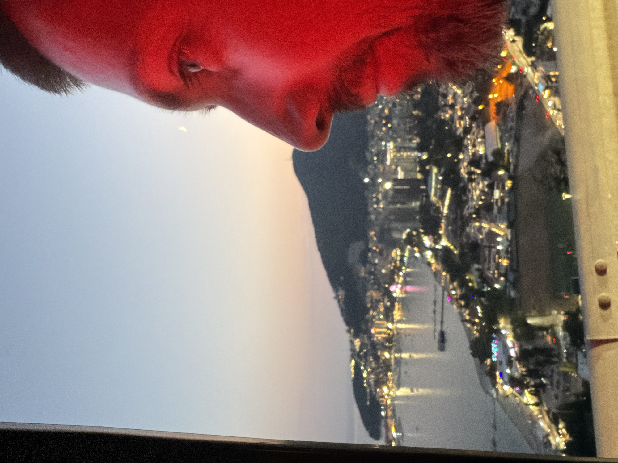 A close-up, profile shot shows the right side of a man's face, illuminated by a red light. He has dark hair, a beard, and is looking towards a city skyline at dusk. The city, identified as Košljun, Montenegro, is spread across a bay and hillside, with numerous lights twinkling from buildings and along the coast. The bay is dotted with boats, and the lights reflect on the water. A faint crescent moon is visible in the pale twilight sky. The foreground of the image features a blurry railing, suggesting the viewpoint is from a balcony, observation deck, or similar elevated structure. The overall impression is one of quiet contemplation against a vibrant urban backdrop.